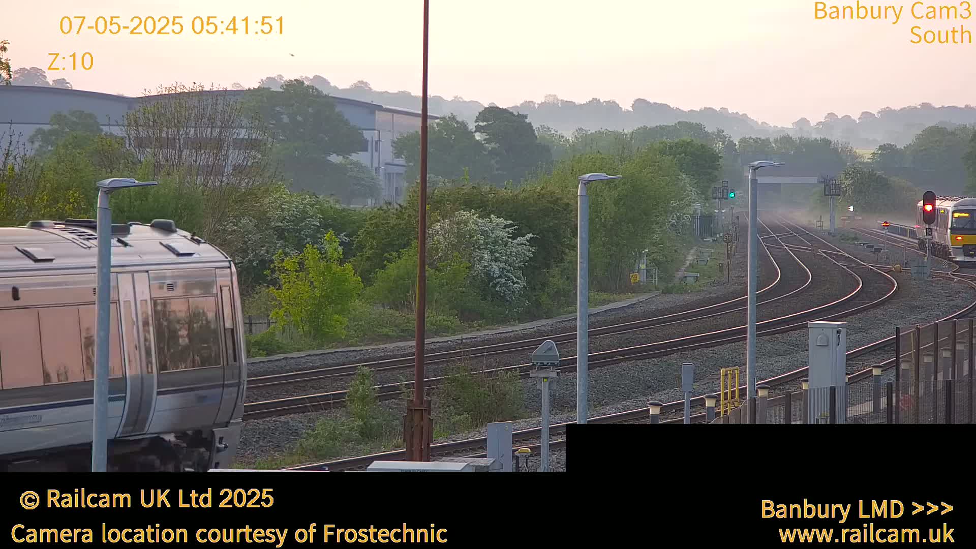 Two trains pass each other on a railway line amidst a hazy dawn, surrounded by greenery and industrial buildings.
