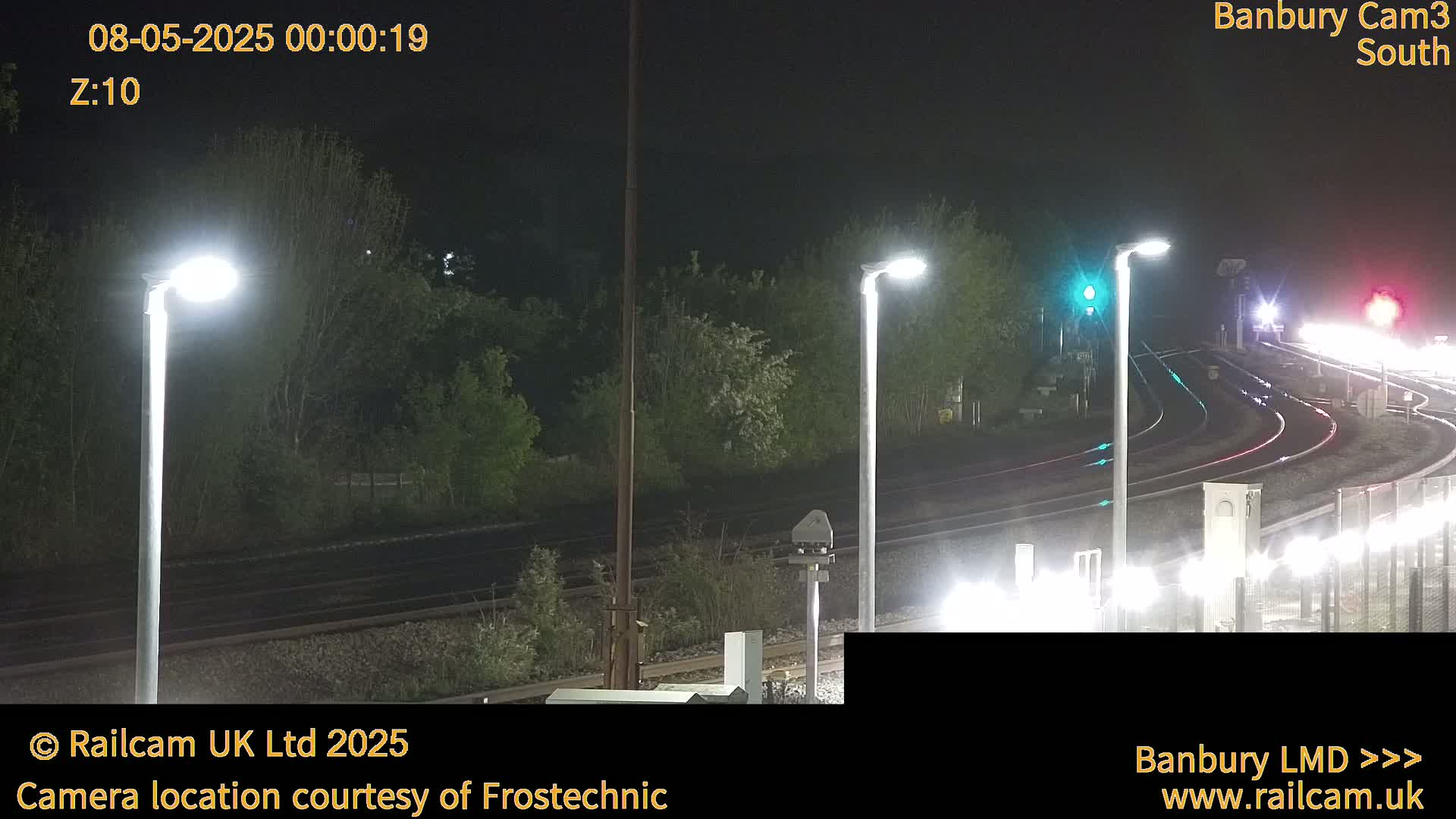 At night, under bright lights, train tracks curve through a landscape of dark green trees and shrubs.