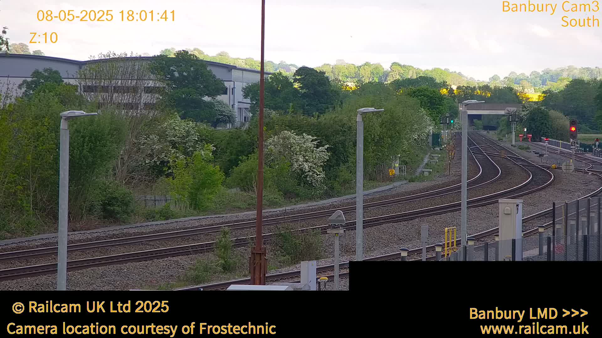 Under a partly cloudy sky, several sets of train tracks curve through a landscape of green vegetation and industrial buildings.