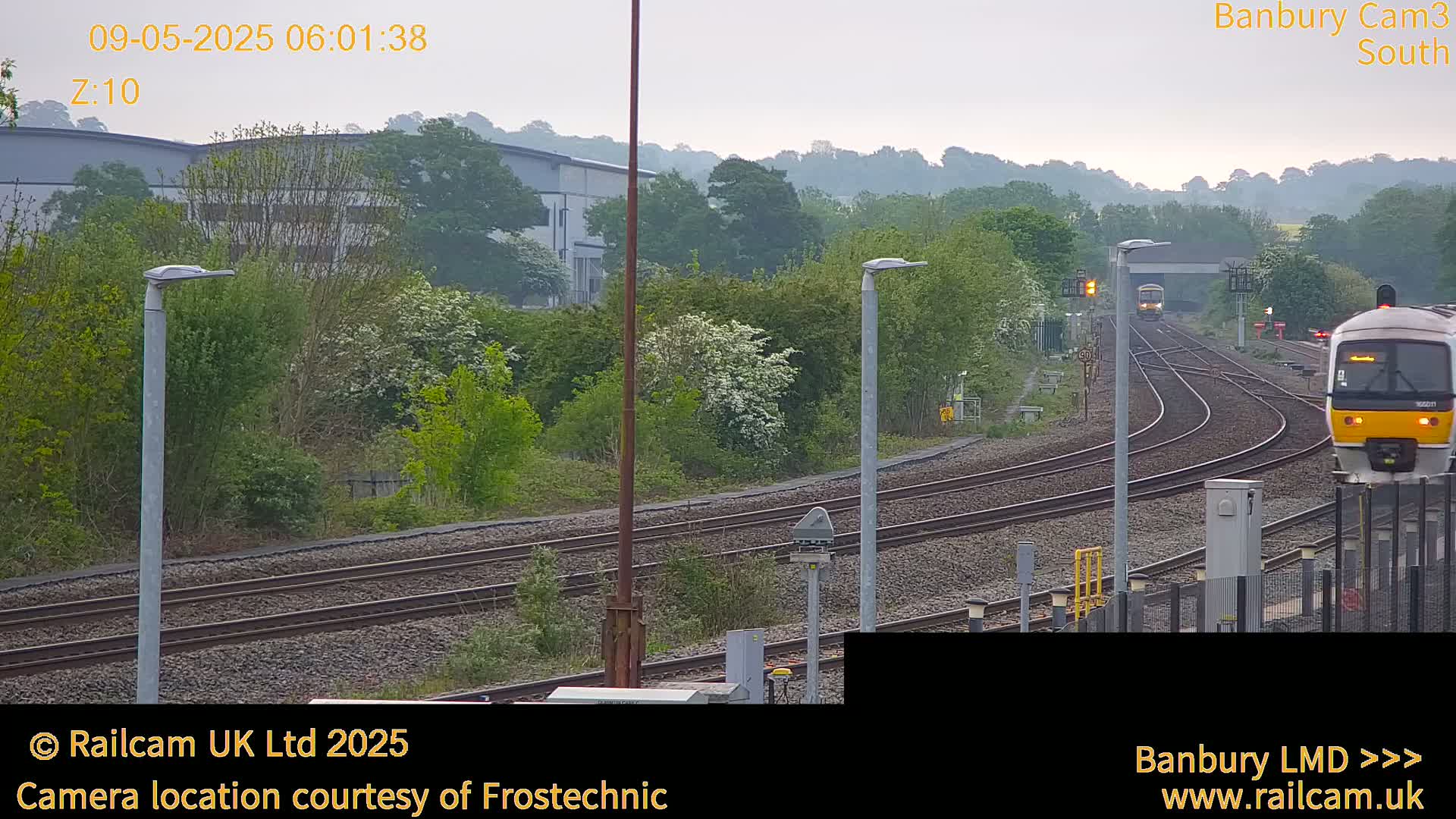 Two trains are visible on a railway line amidst a landscape of green trees and shrubs under an overcast sky.