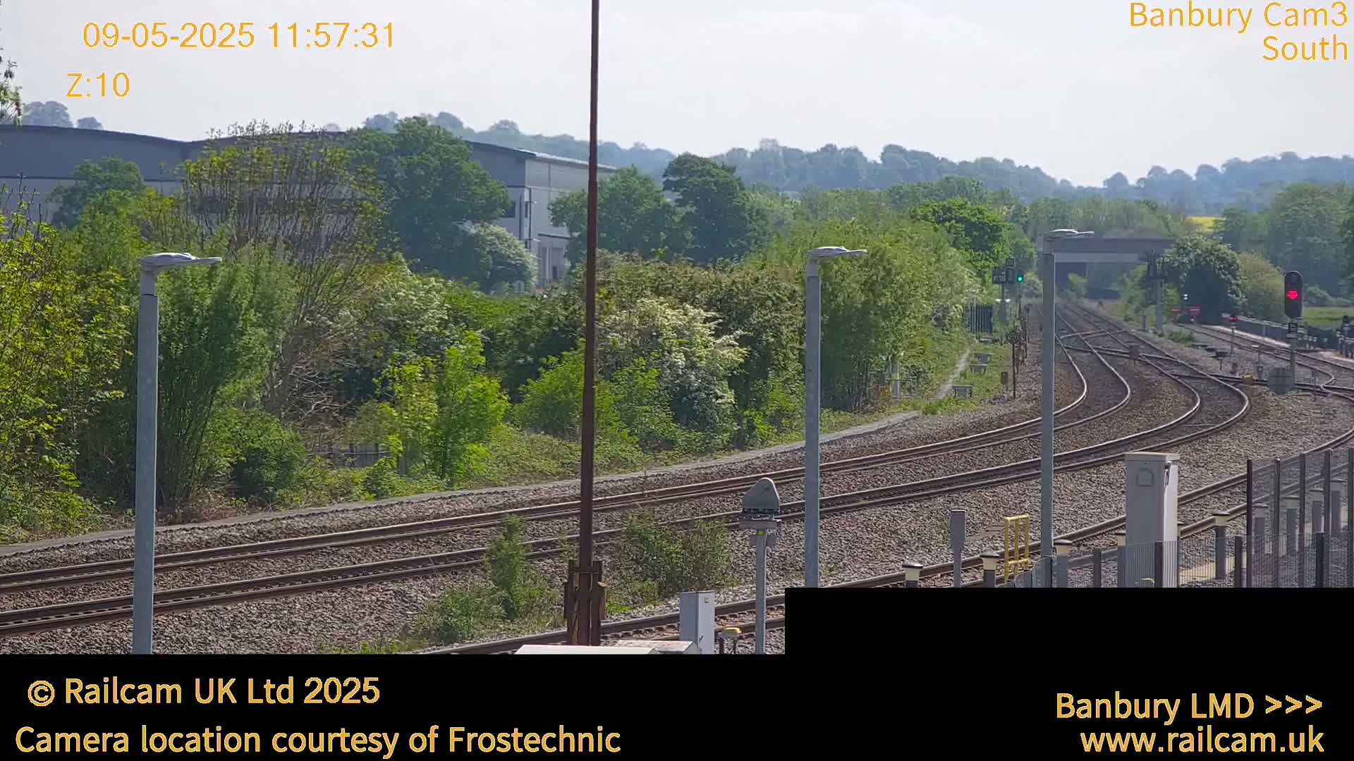 Under a sunny sky, multiple train tracks curve through a landscape of green trees and shrubs, near a building.