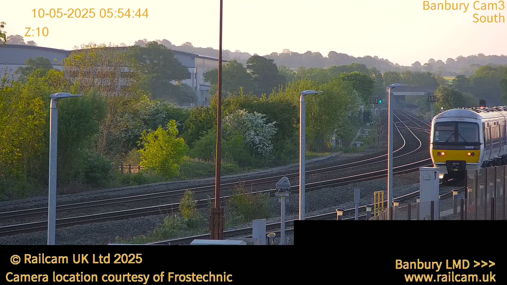 A train approaches a station on a slightly hazy morning, passing alongside leafy trees and industrial buildings.
