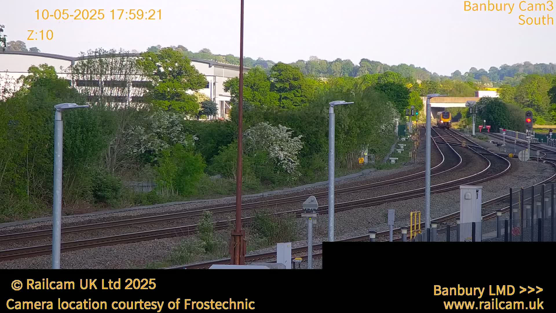A train passes through a railway station on a partly sunny day, surrounded by lush green vegetation and industrial buildings.