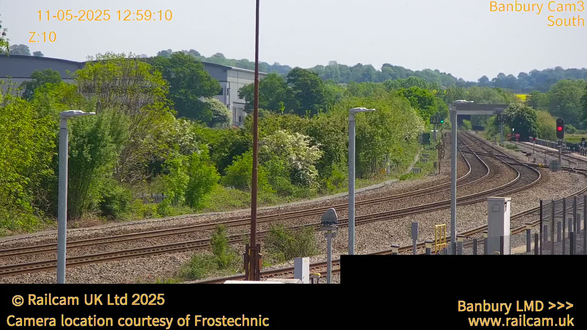 A sunny view of several train tracks curving through a lush green landscape, with buildings and trees in the background.