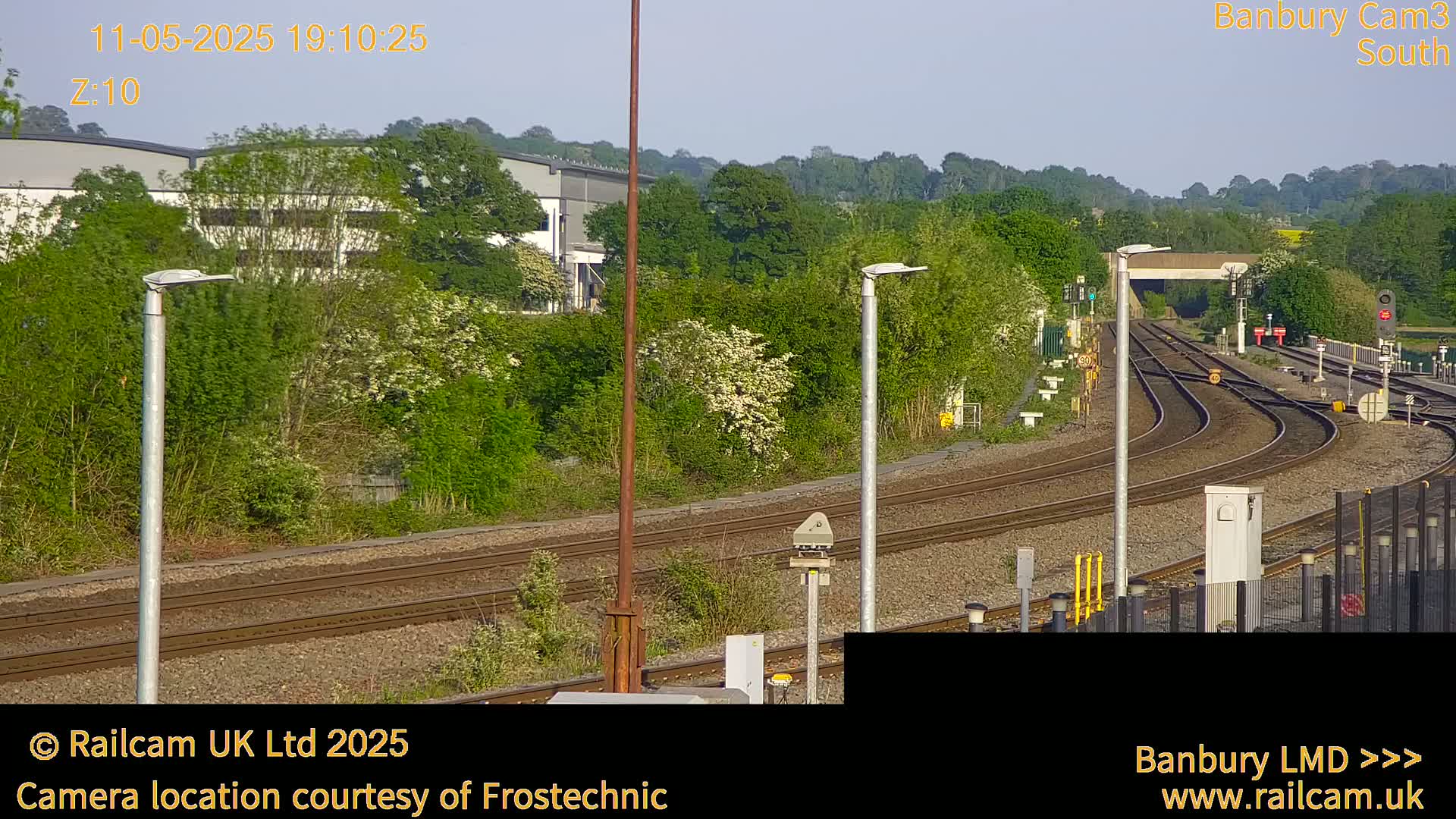 Under a bright, clear sky, multiple train tracks curve through a landscape of green trees and bushes, alongside industrial buildings.