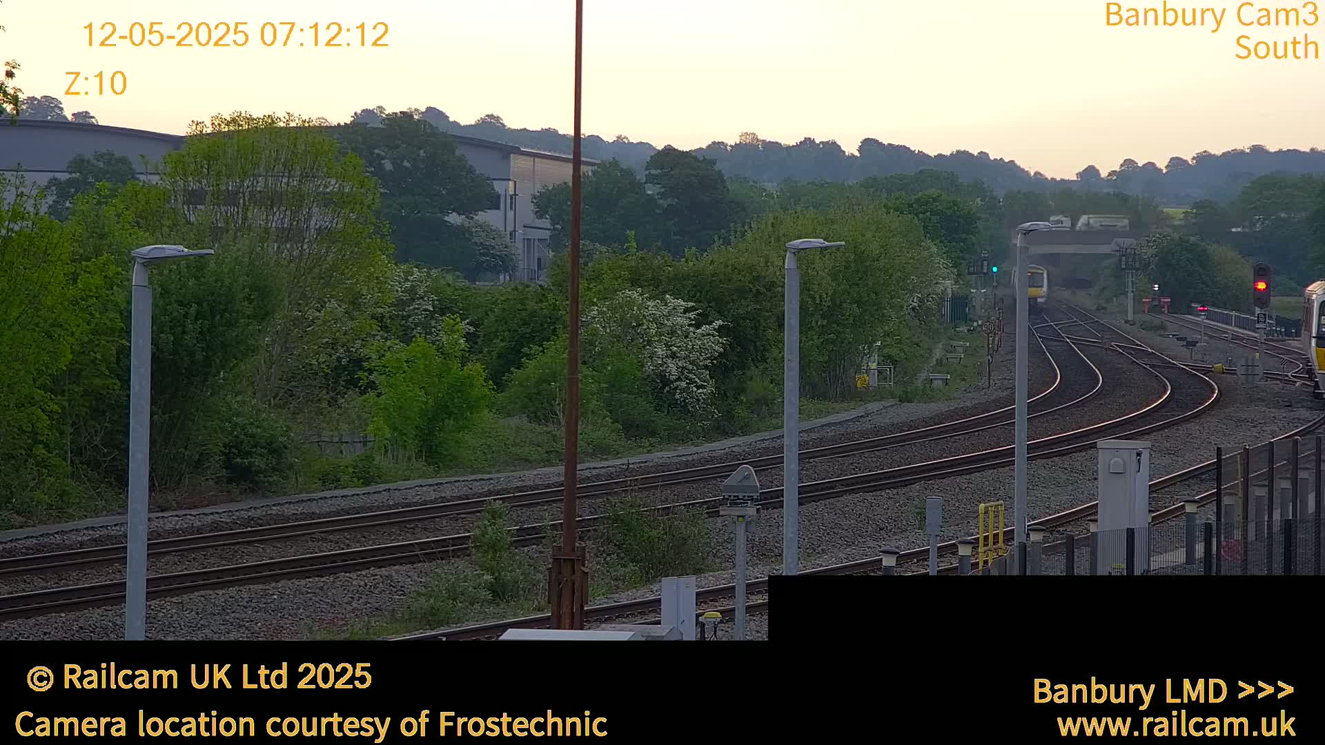 A train approaches a railway junction at dawn under a mostly clear sky, surrounded by green vegetation and industrial buildings.