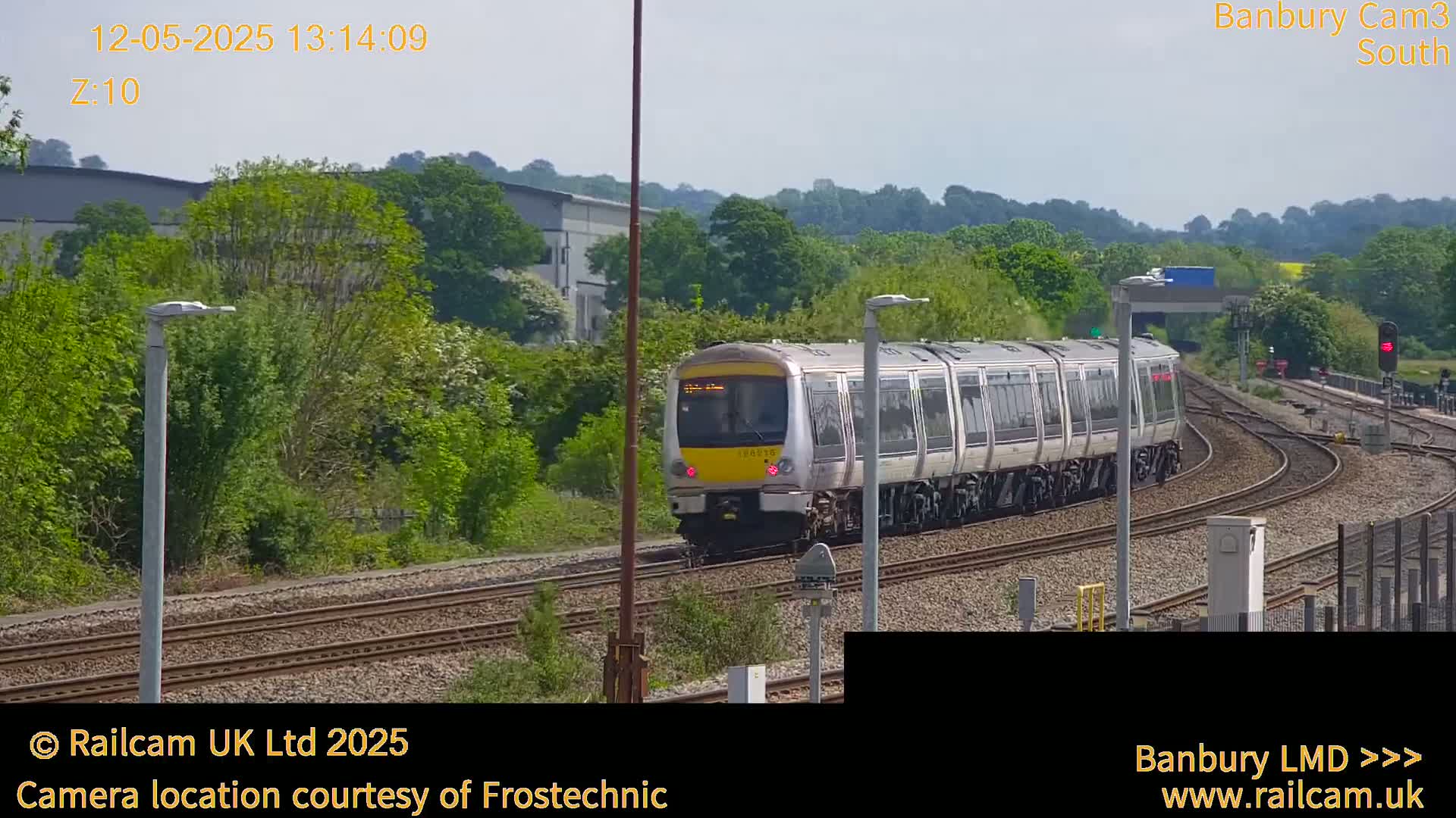 A long, light-grey passenger train moves along a railway track on a partly sunny day, passing through a landscape of green trees and industrial buildings.