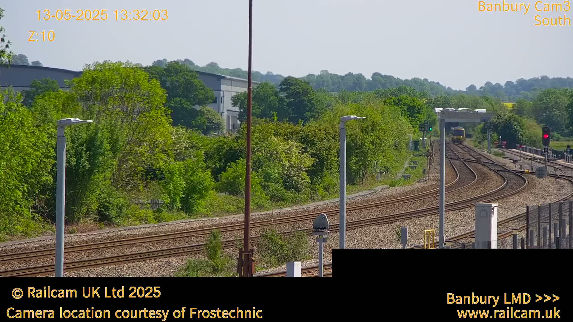 A yellow train approaches a railway junction on a sunny day, surrounded by lush green trees and vegetation.