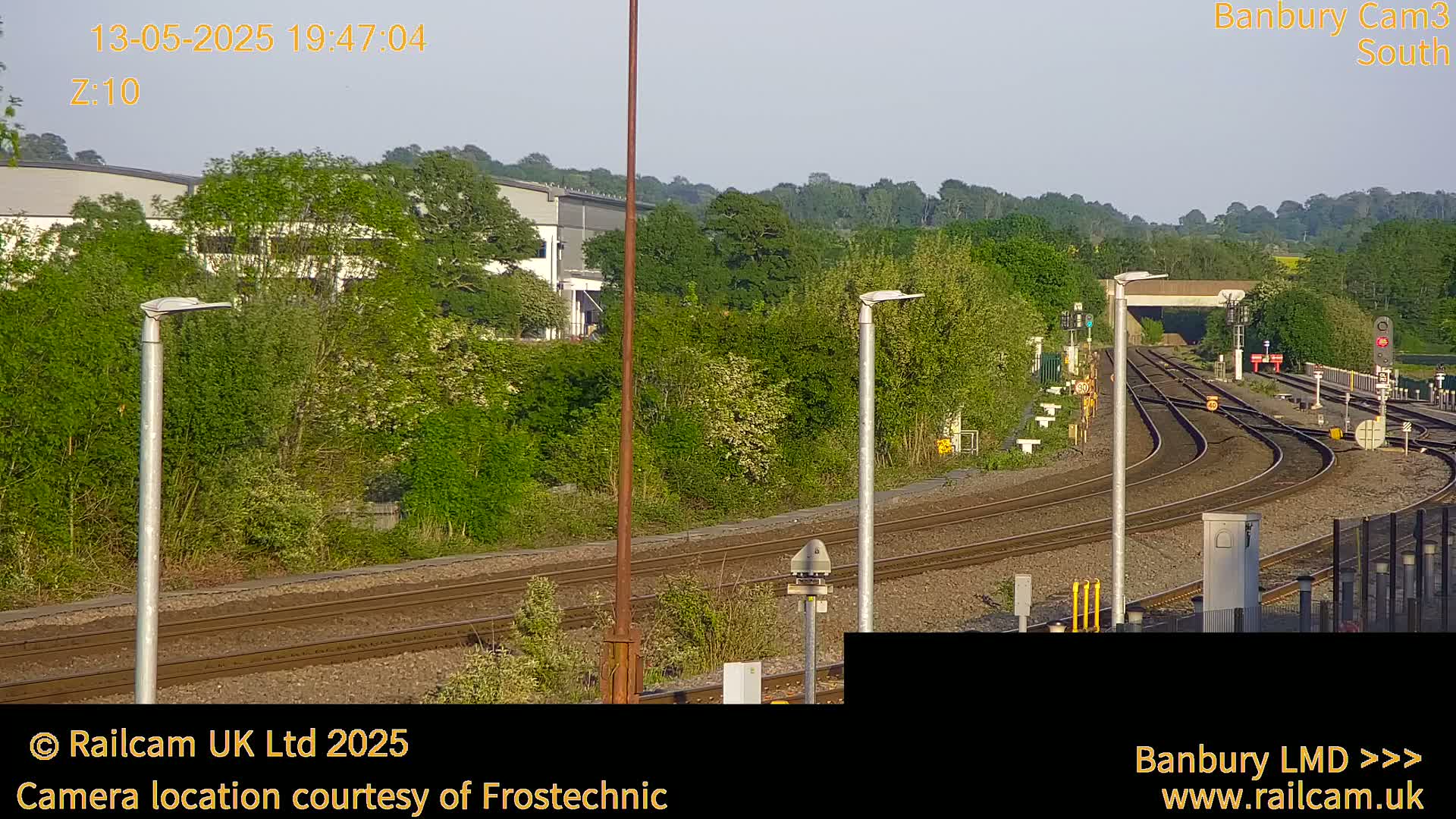 Several sets of train tracks curve through a lush green landscape under a clear sky at dusk, passing near industrial buildings and tall trees.