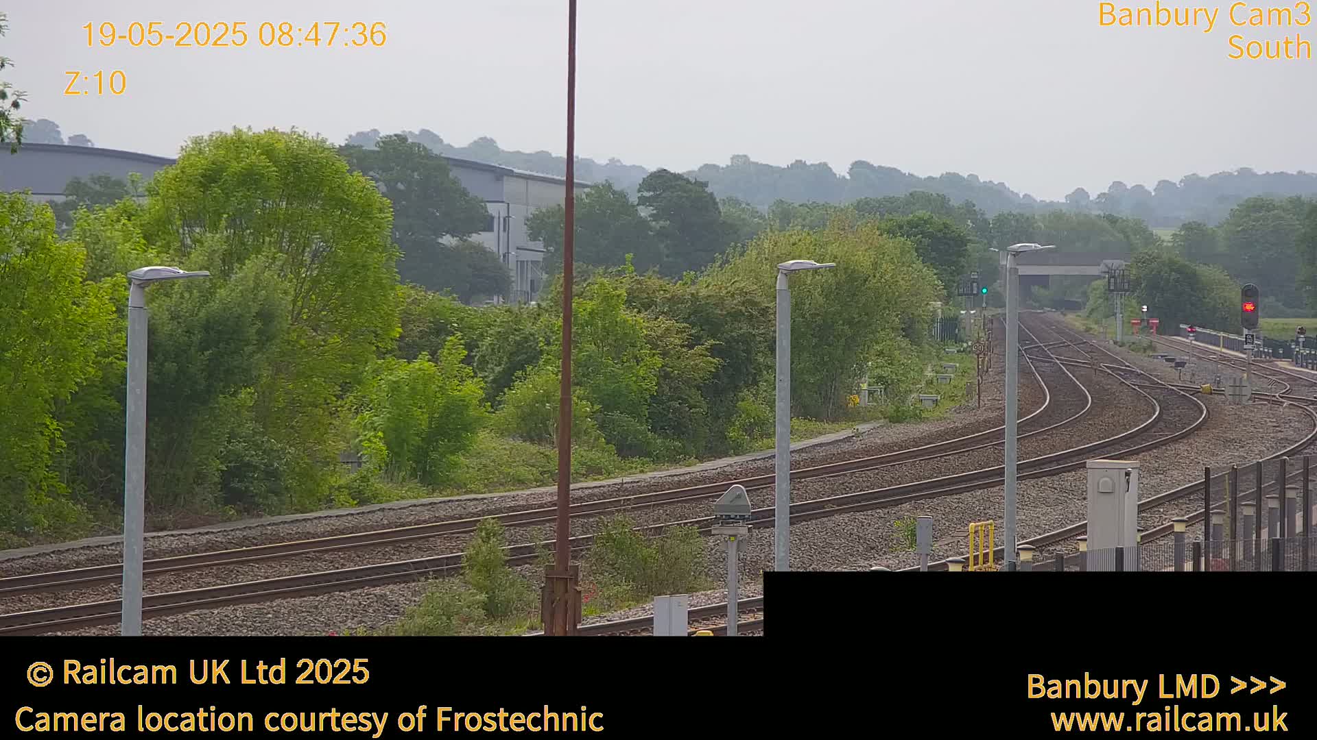 A hazy view of curving train tracks leading to a distant hillside, with lush green vegetation and several lamp posts lining the railway.