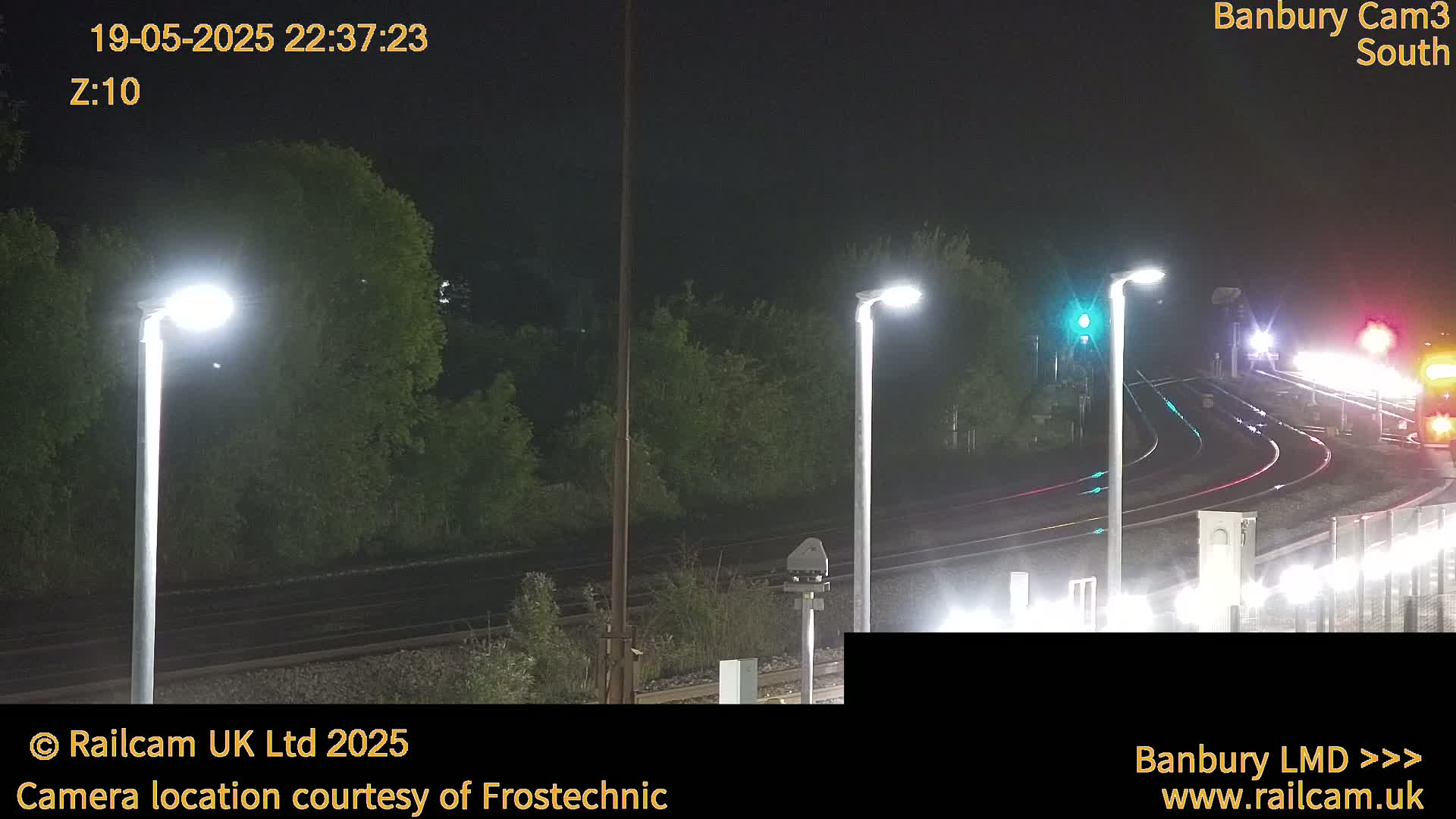 A nighttime view of railroad tracks and train lights, illuminated by streetlights, with surrounding trees and vegetation.