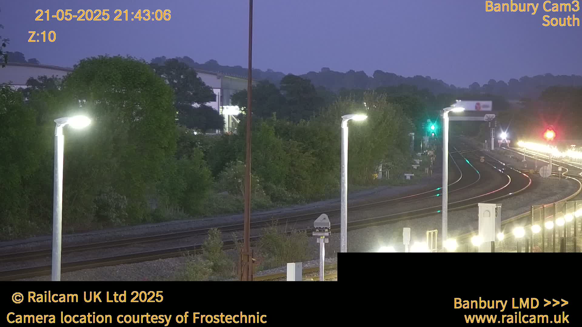 Under a twilight sky, illuminated train tracks curve through a landscape of trees and buildings, with several lit lampposts lining the tracks.