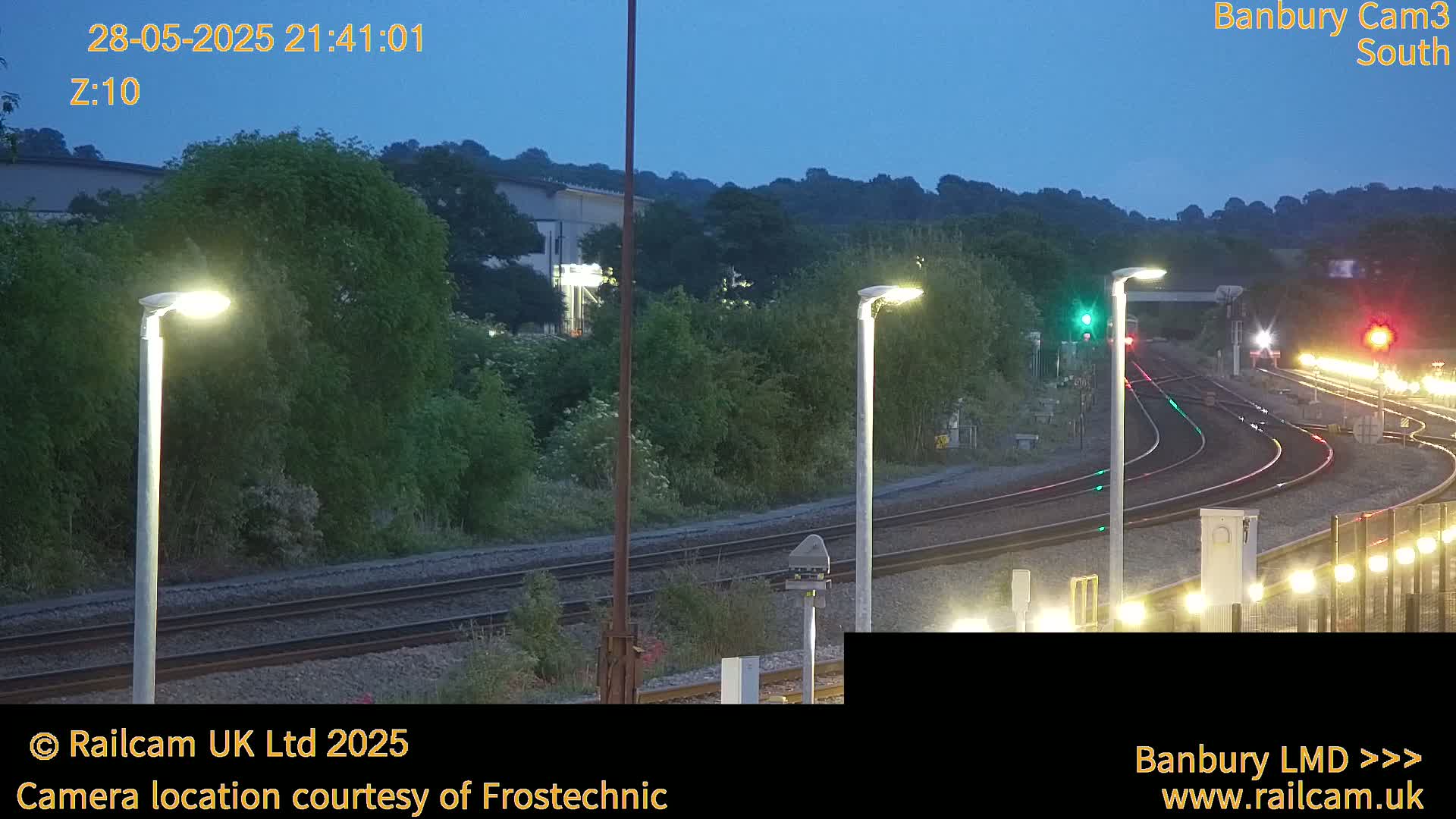 Under a twilight sky, illuminated train tracks curve through a landscape of green vegetation and lit lamp posts.
