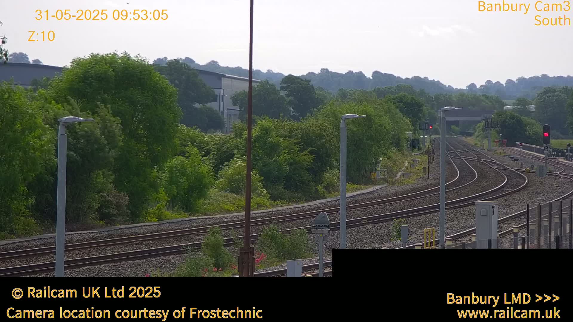 A view of a railway yard with multiple train tracks curving through a landscape of green trees and shrubs under a hazy sky.
