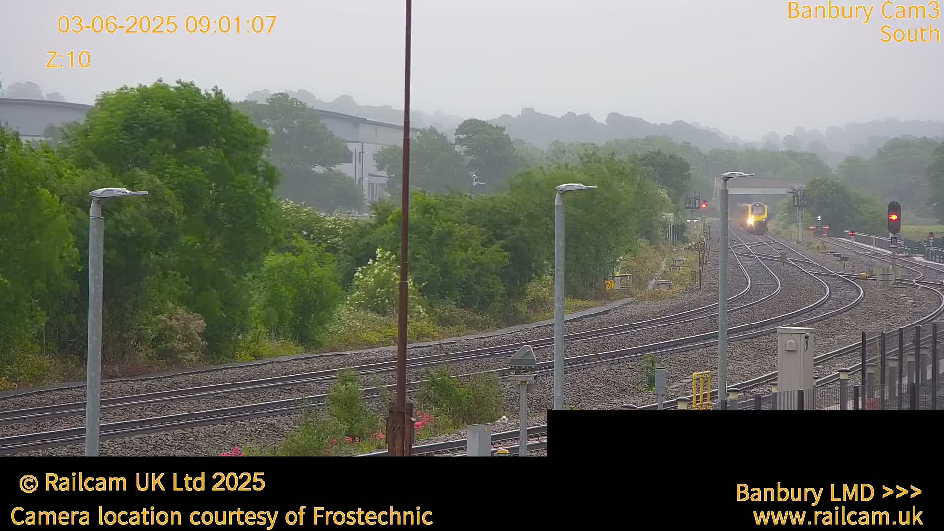 A yellow train approaches a railway station on a foggy, overcast day.