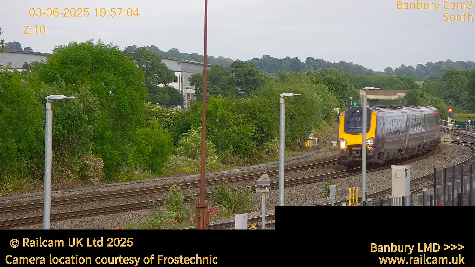 A gray and yellow passenger train moves along railway tracks past lush green vegetation under an overcast sky.