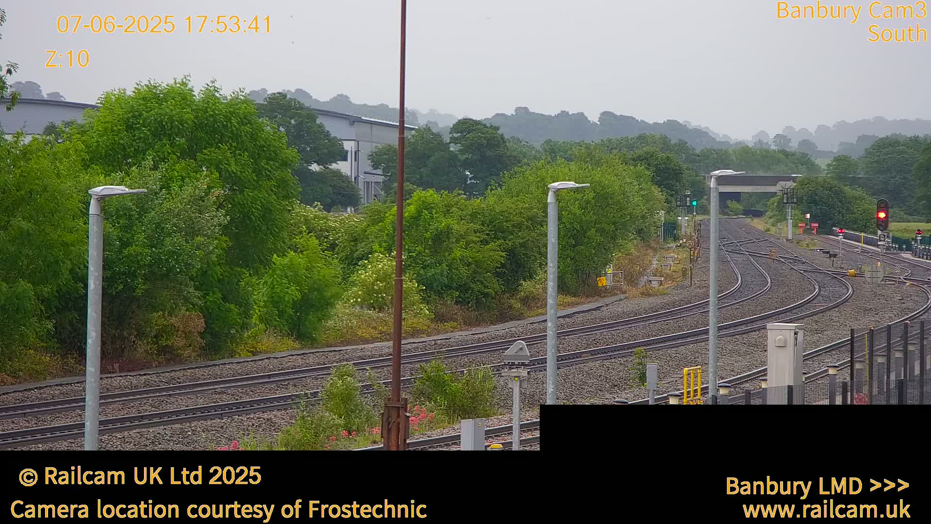 A view of a railway track system on an overcast day, surrounded by lush green vegetation and some industrial buildings in the background.