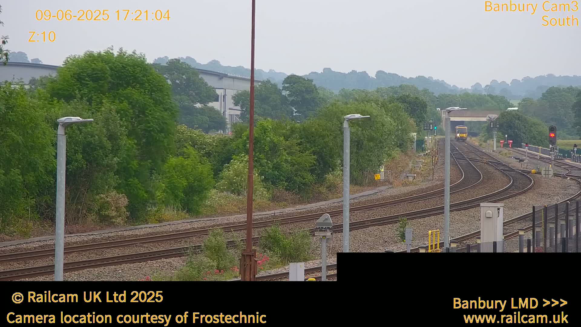 A train is seen approaching a railway station on a slightly overcast day, surrounded by greenery and some industrial buildings in the distance.