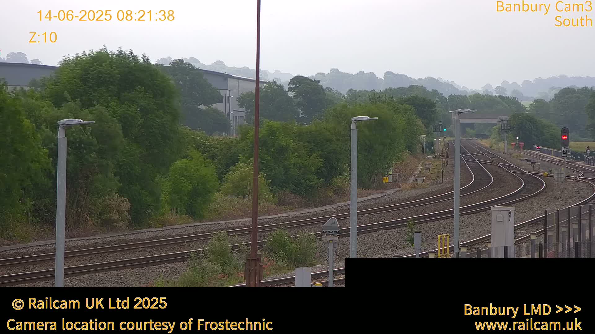 A hazy view of a train yard with multiple tracks curving through a landscape of green trees and shrubs under an overcast sky.