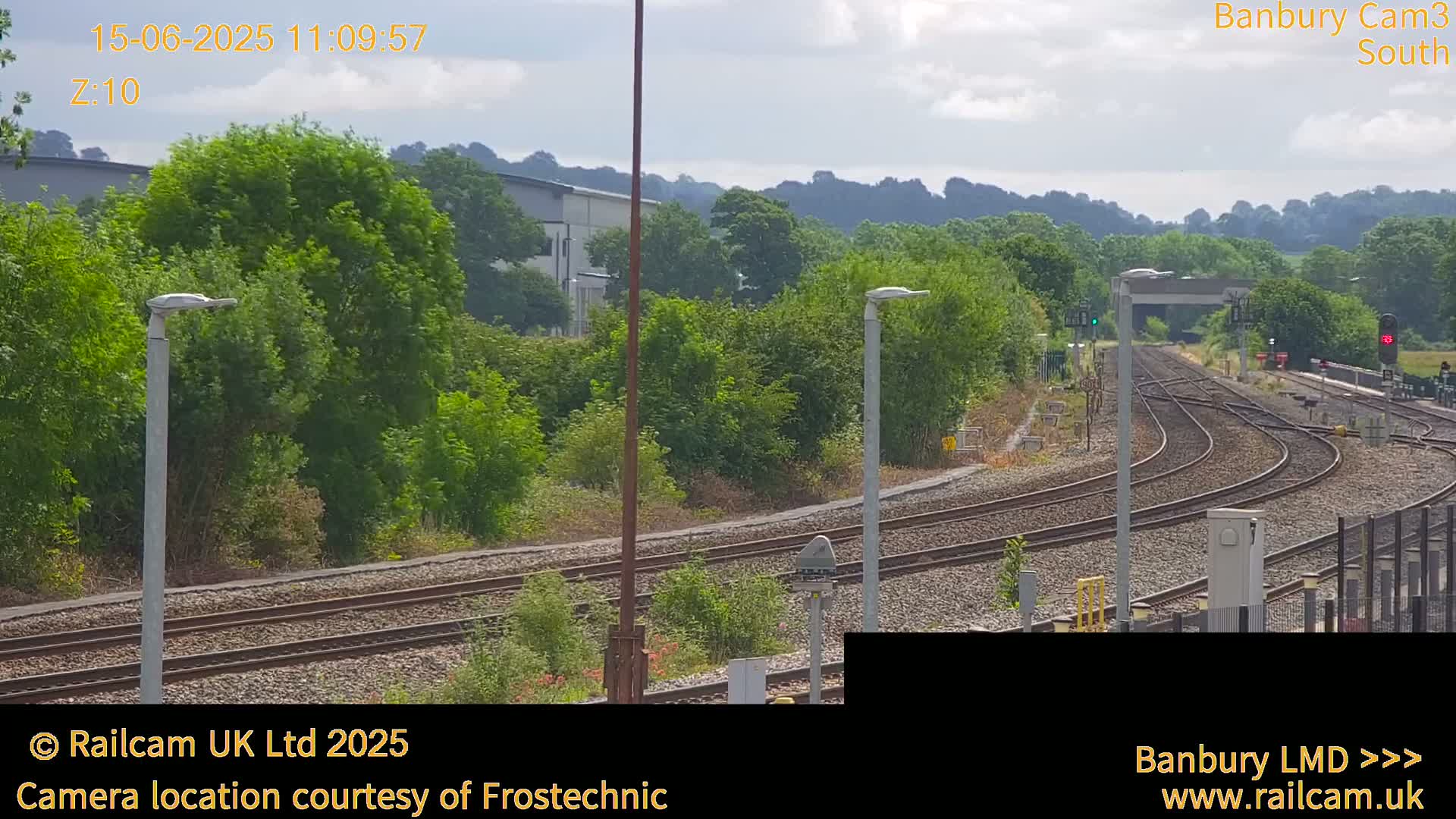 A sunny view of a railway yard with multiple tracks curving through lush green vegetation under a partly cloudy sky.