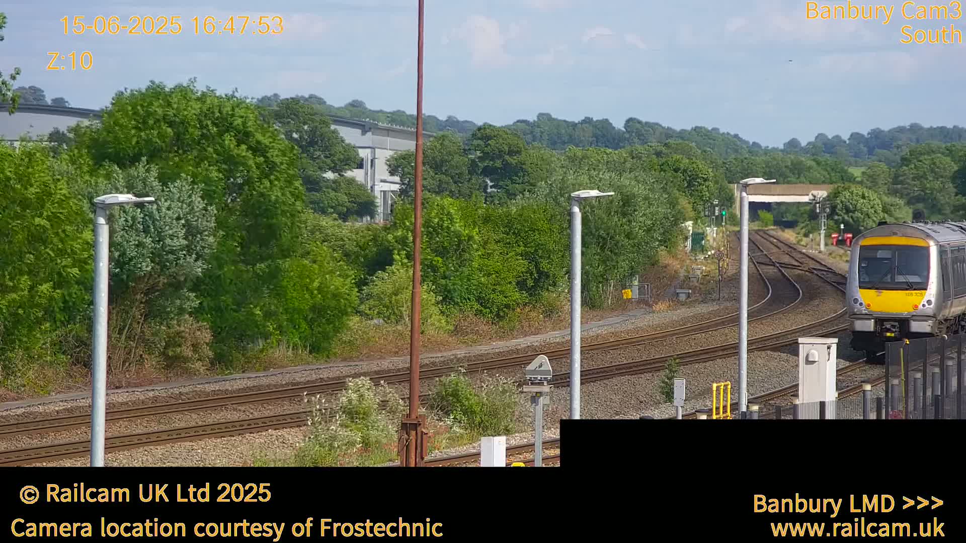 A train approaches a railway station on a sunny day, surrounded by green vegetation and industrial buildings in the distance.