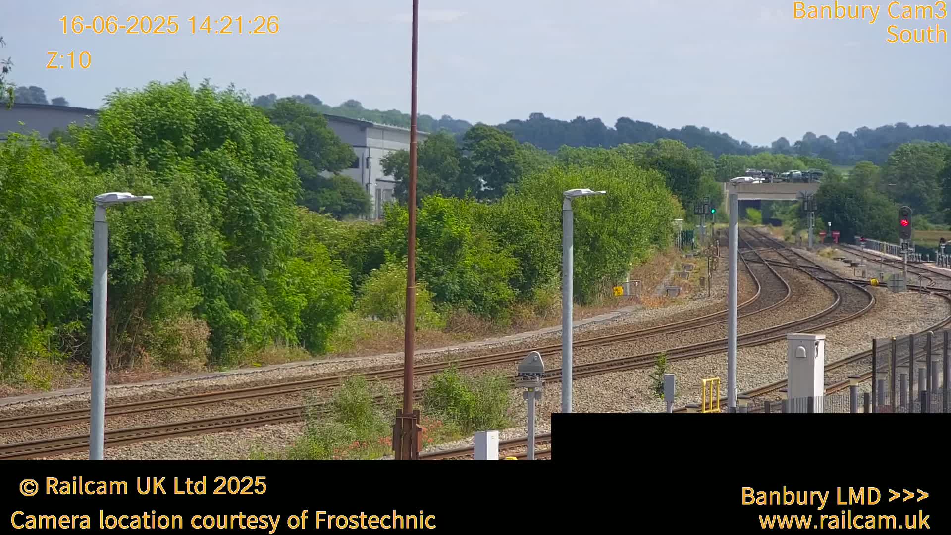 A sunny outdoor view of a railway line curving through a landscape of green trees and shrubs, with several lampposts along the track.
