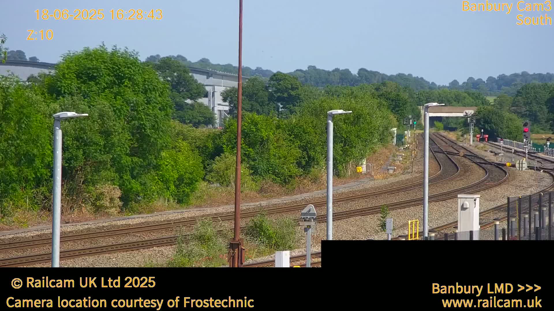 A sunny view of a railway track curving through a green landscape, with several tall lampposts and trees alongside.