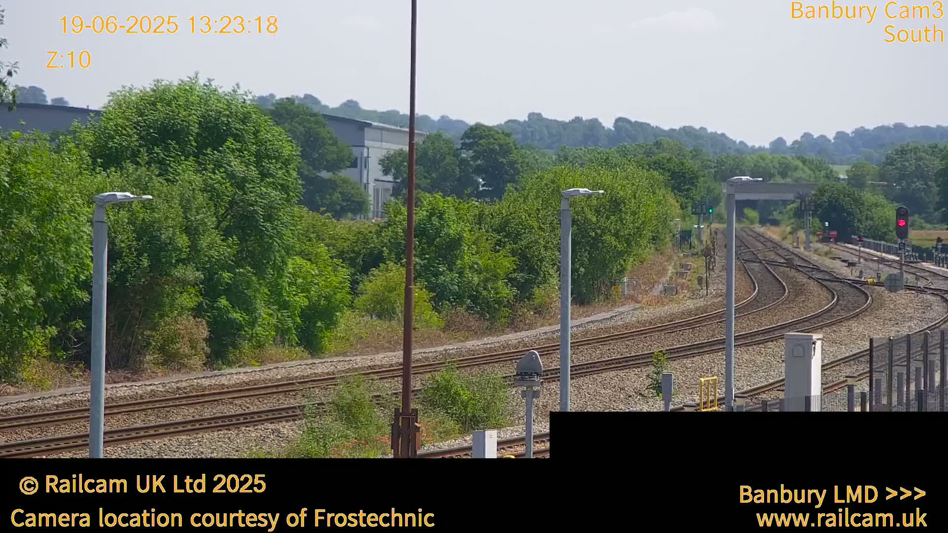 A sunny view of several train tracks curving through a landscape of green trees and shrubs, with lampposts along the tracks and a building in the distance.
