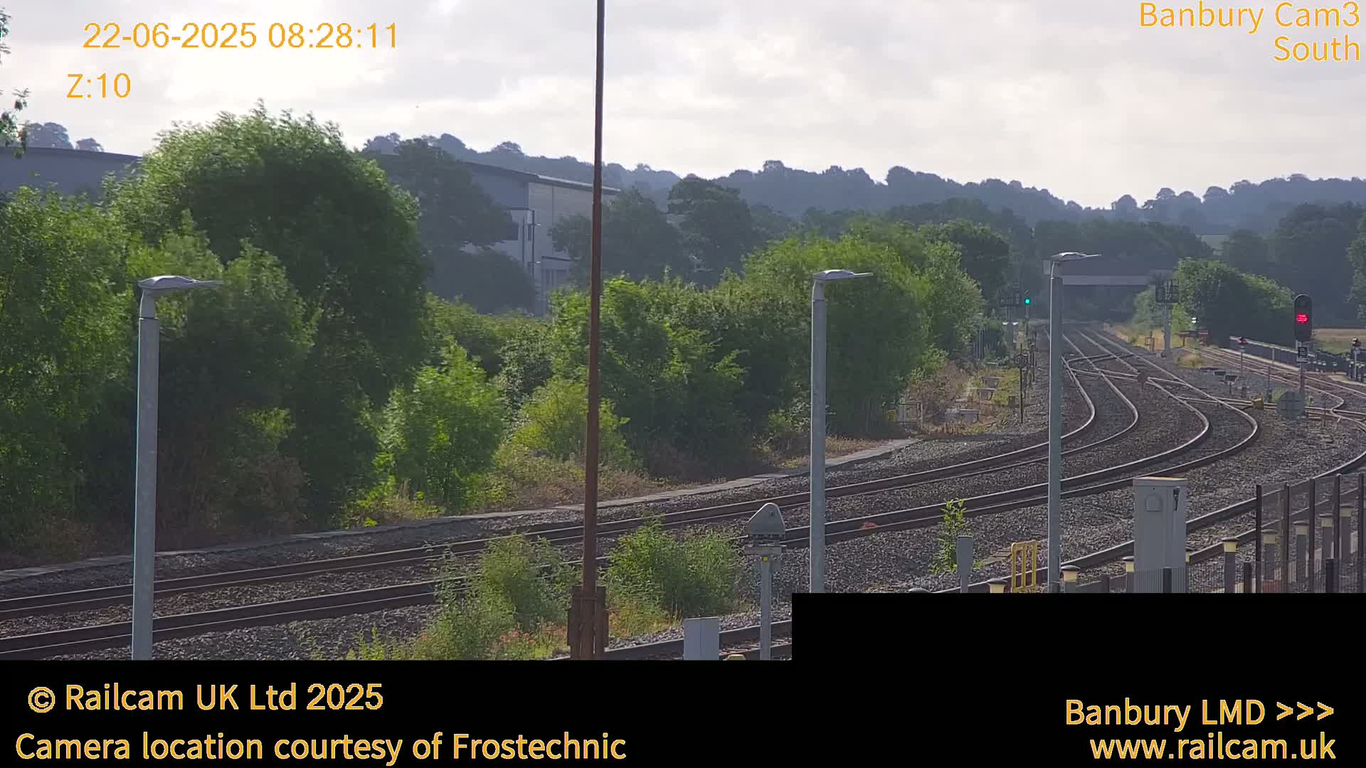 A slightly overcast day shows a view of several train tracks curving through a lush green landscape with trees and tall light posts.