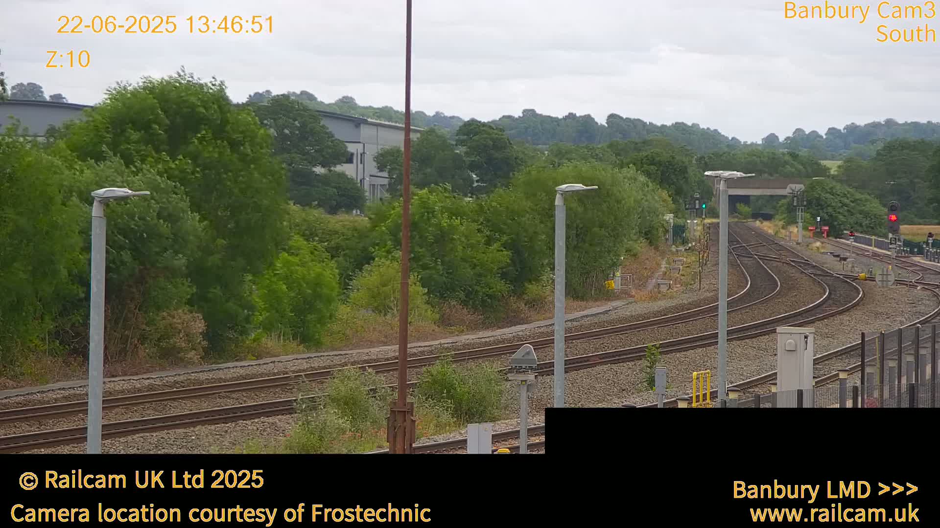 A view of curving train tracks on an overcast day, surrounded by lush green trees and industrial buildings in the distance.