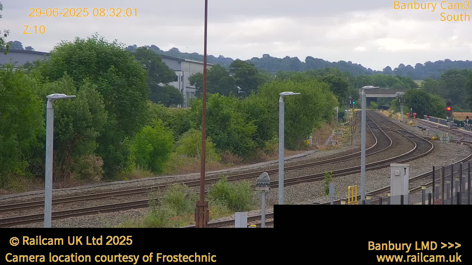 A view of curving train tracks leading to a railway station under an overcast sky, surrounded by green vegetation and buildings in the distance.