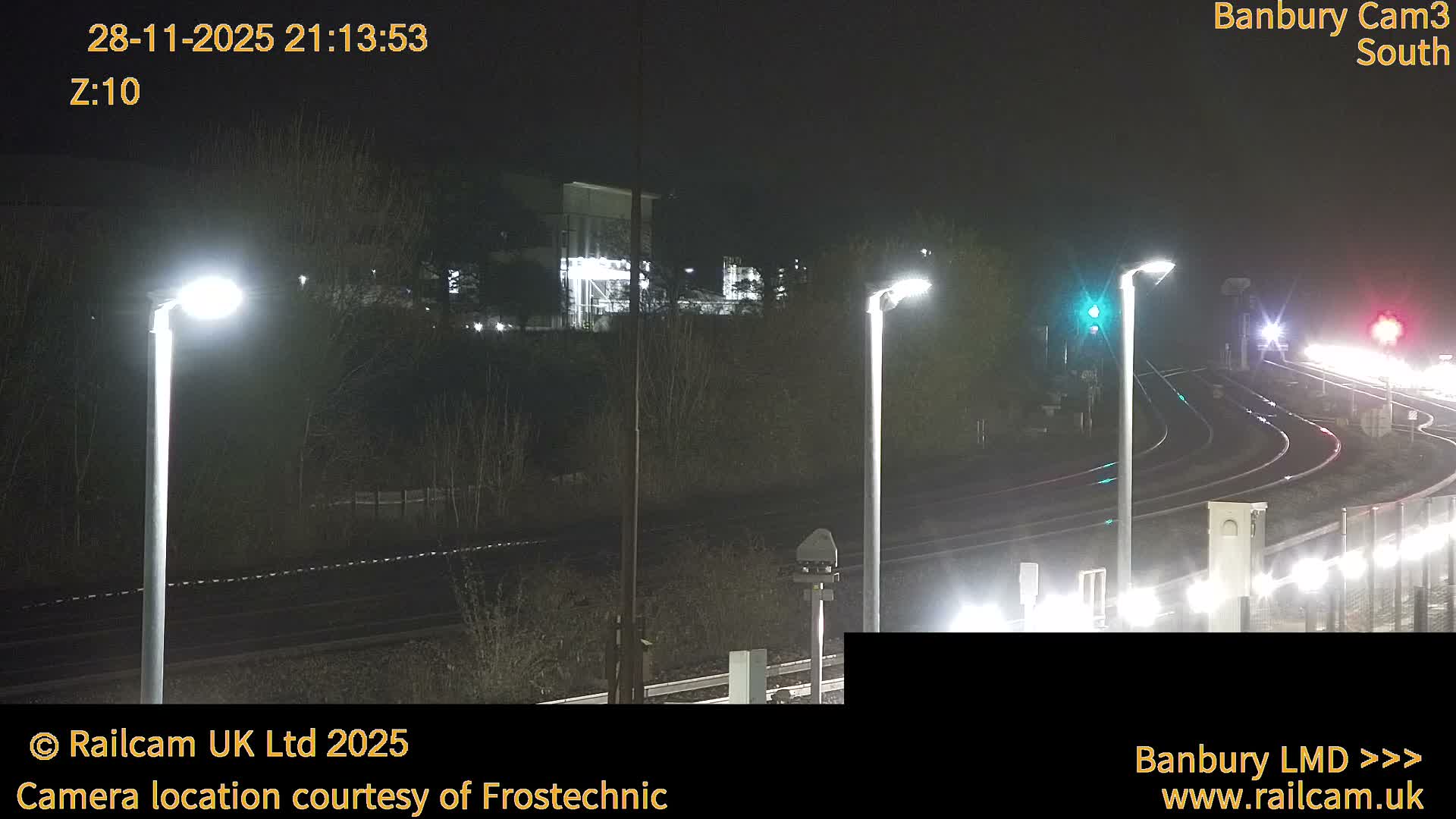 A nighttime image captures a rainy railway scene with multiple tracks, bright streetlights, a green signal, and a distant red signal, all reflected vividly on the wet ground.