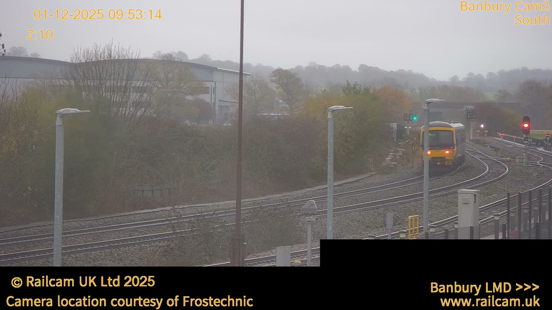 A yellow and grey train approaches on multiple railway tracks with signals displaying green and red, through a misty landscape of bare trees and buildings under a grey, overcast sky.