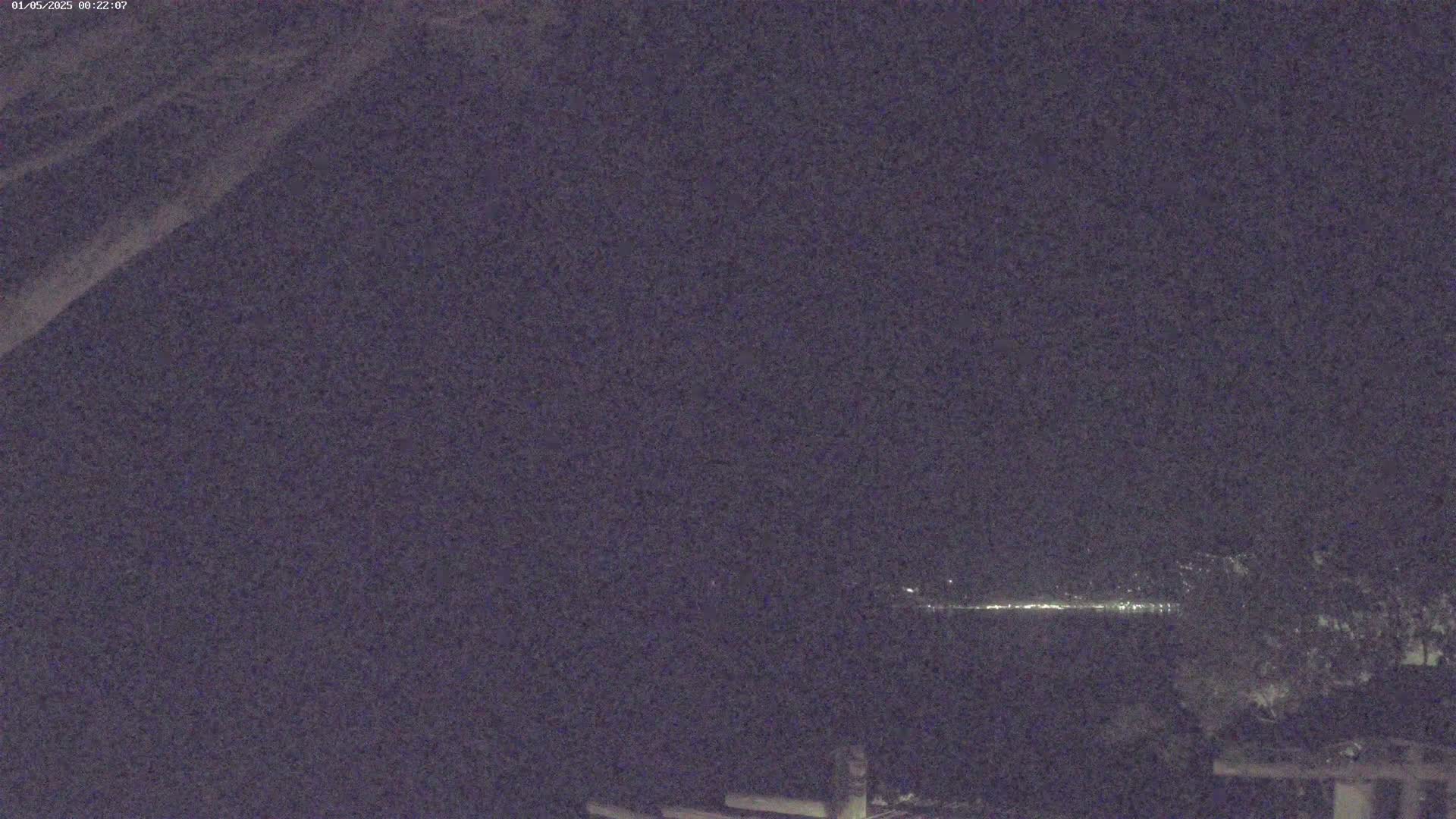 A nighttime, low-light view shows a distant line of lights across a dark body of water, with some dark vegetation in the foreground and a portion of what appears to be a structure at the bottom.