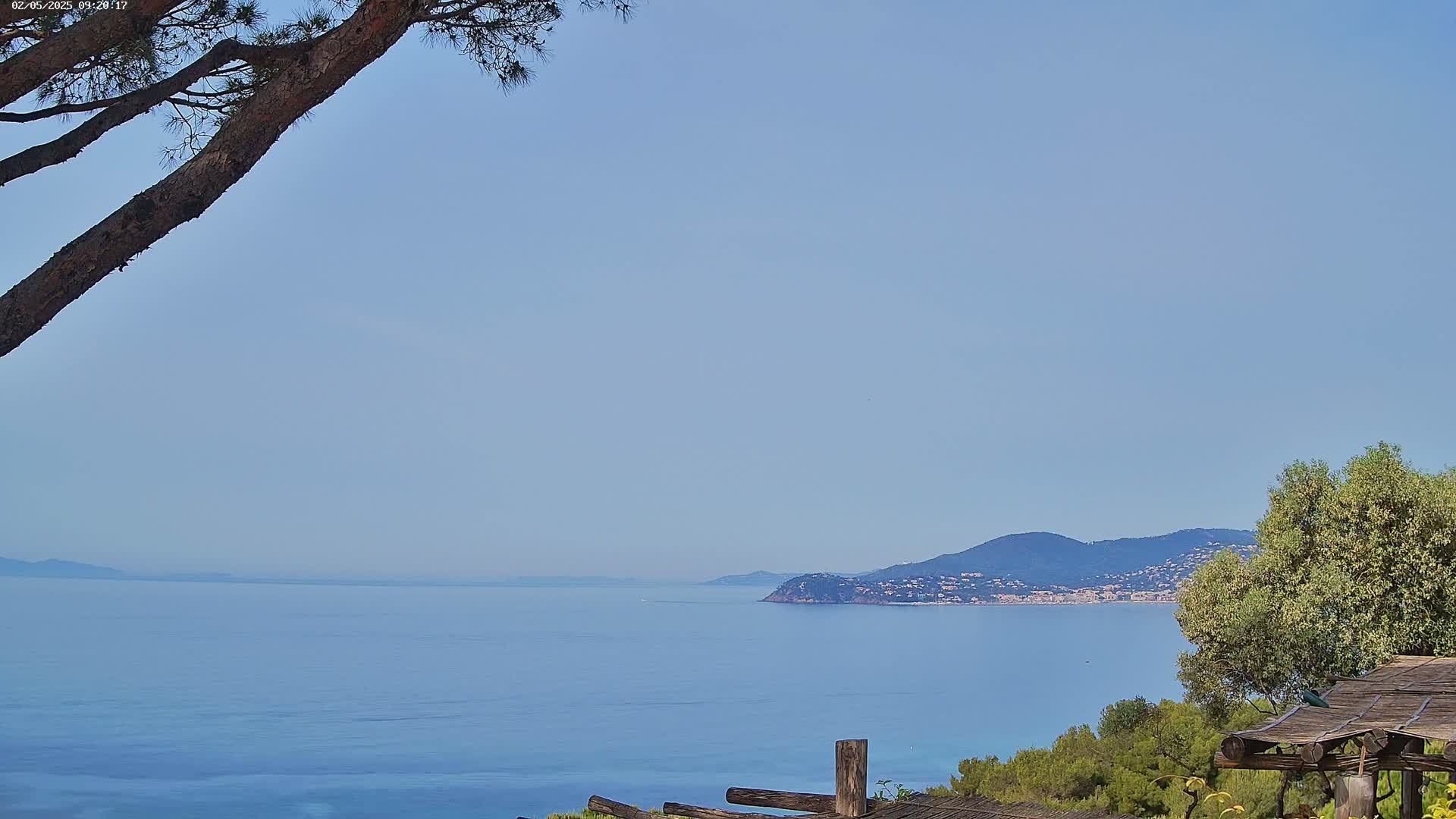 A tranquil coastal scene on a clear, sunny day features a calm blue sea stretching to a distant mountainous shoreline, partially obscured by a tree branch in the foreground and a wooden structure.