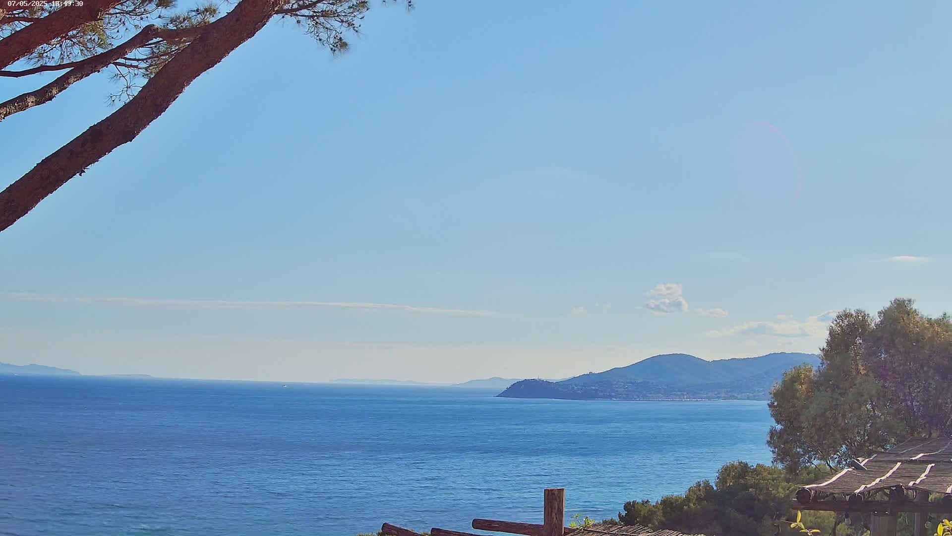 A tranquil ocean scene under a clear blue sky shows a distant mountain range, partially obscured by a tree in the foreground.