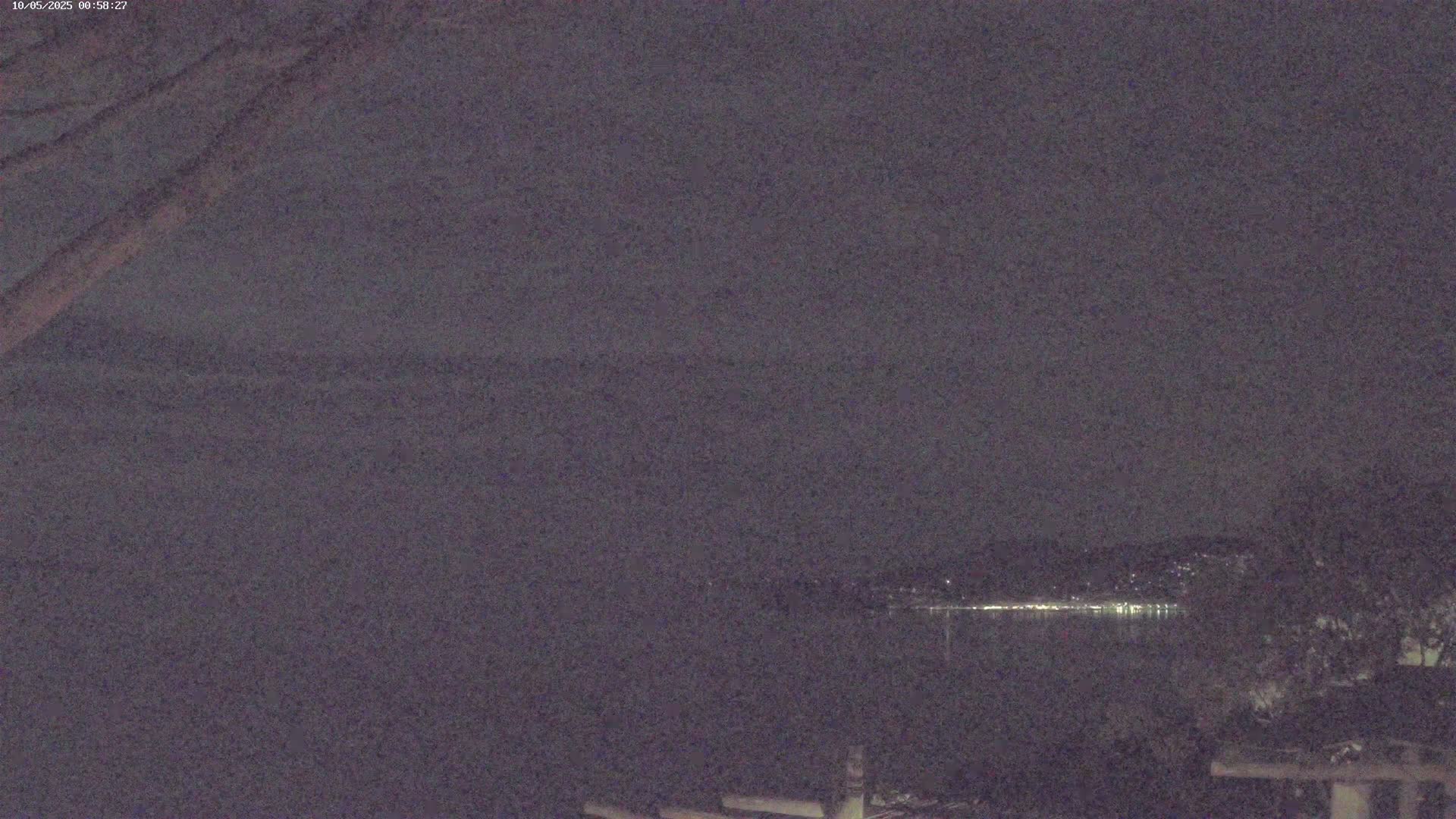 A nighttime view of a dark body of water with a faintly lit shoreline in the distance under a cloudy sky.