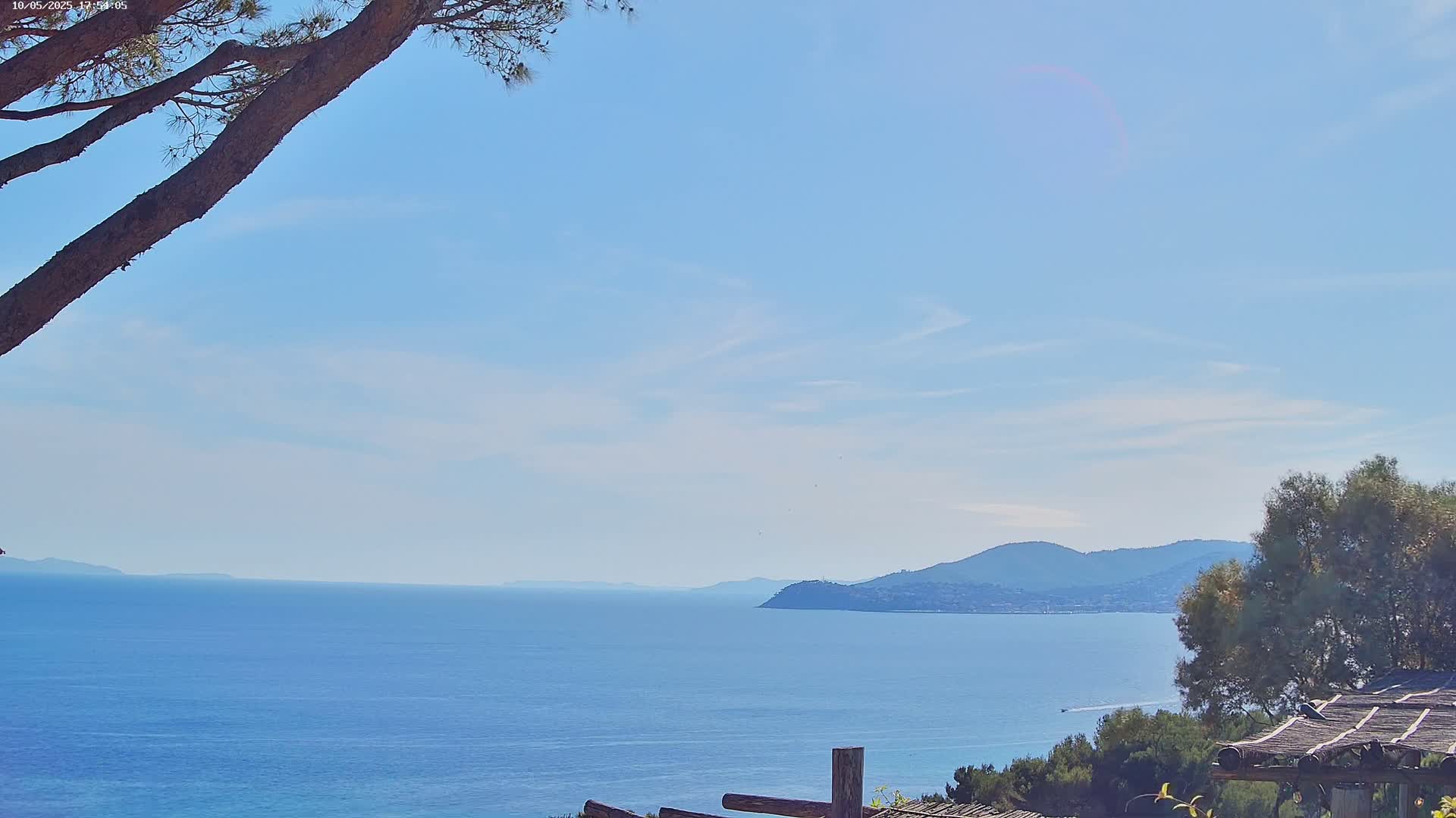 A calm blue ocean stretches to distant hazy mountains under a mostly clear, light blue sky, partially obscured by a tree branch in the foreground and a shaded structure with vegetation.