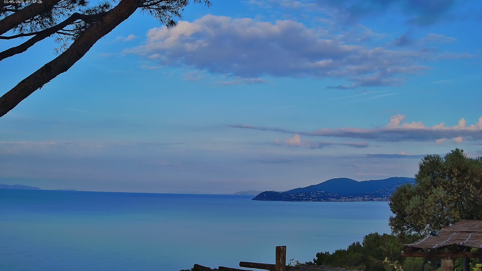 A tranquil coastal scene under a partly cloudy blue sky features a calm, blue sea extending to a distant mountain range, partially obscured by overhanging tree branches.