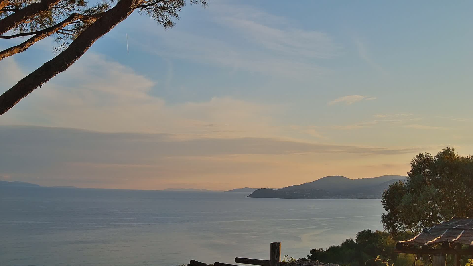 A calm sea stretches to a distant, hilly coastline under a pale, partly cloudy sunset sky.