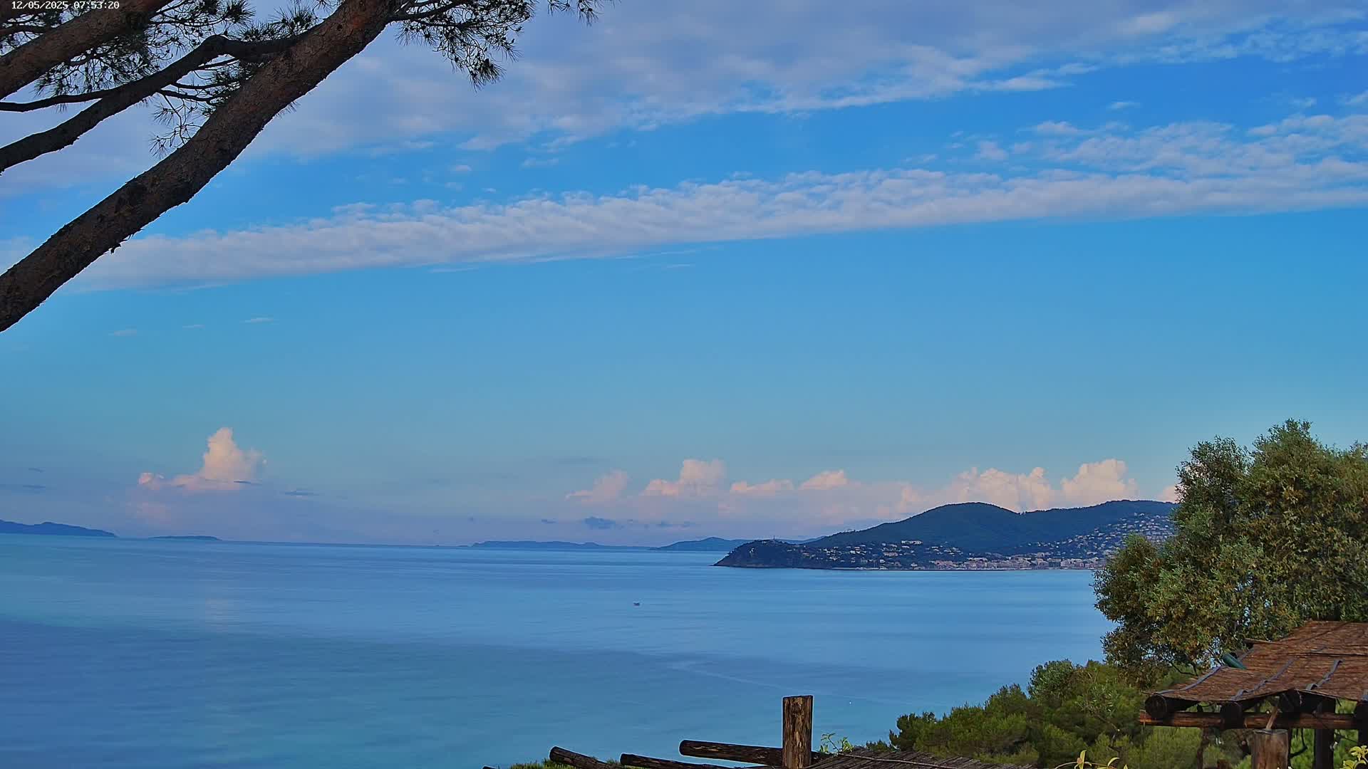 A calm, blue sea stretches to a distant mountainous coastline under a mostly clear, light-blue sky with scattered cumulus clouds.