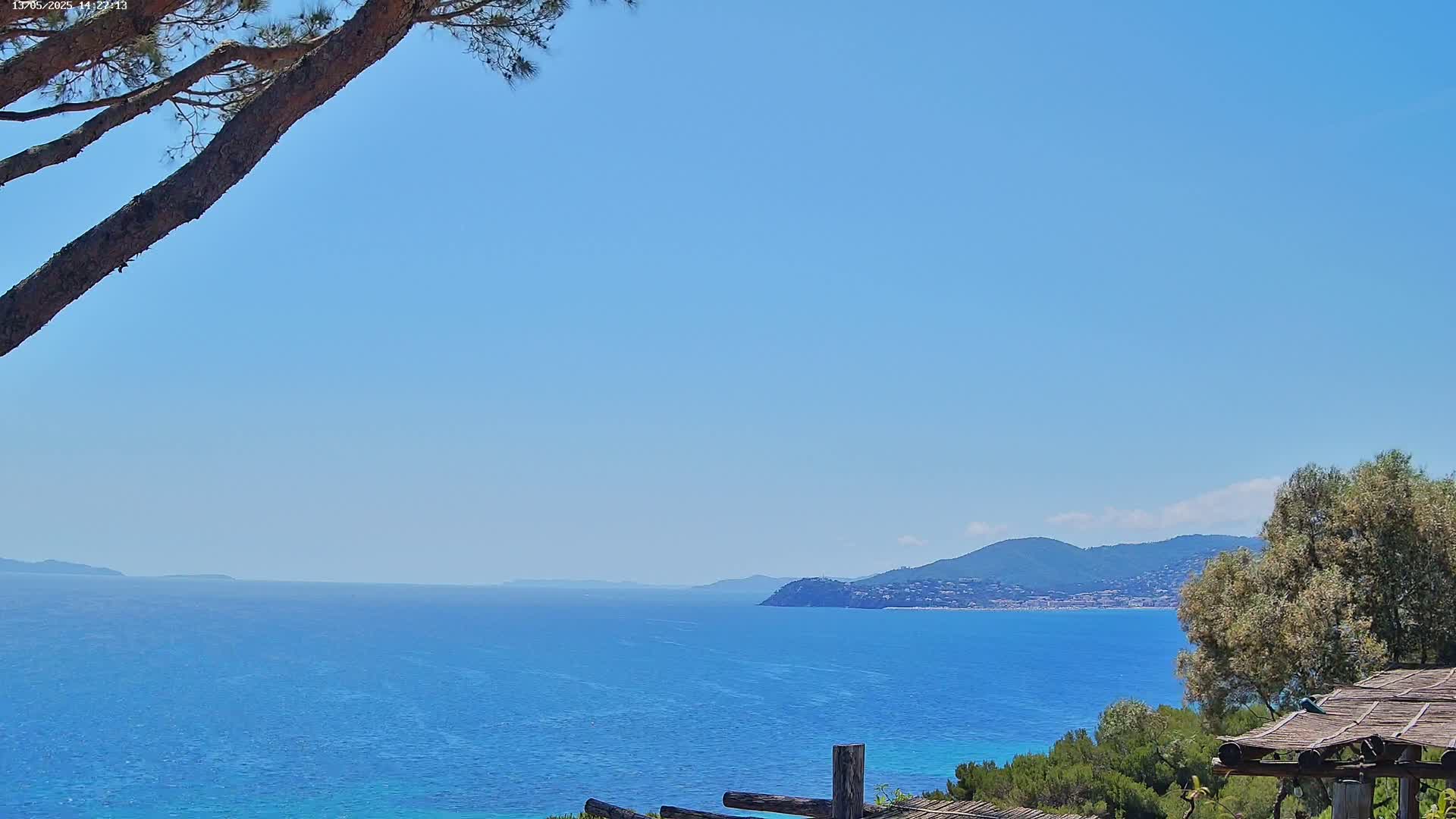 A tranquil coastal scene under a clear blue sky shows a calm blue sea extending to distant mountains, partially obscured by a pine branch in the foreground and some shrubs and a wooden structure.