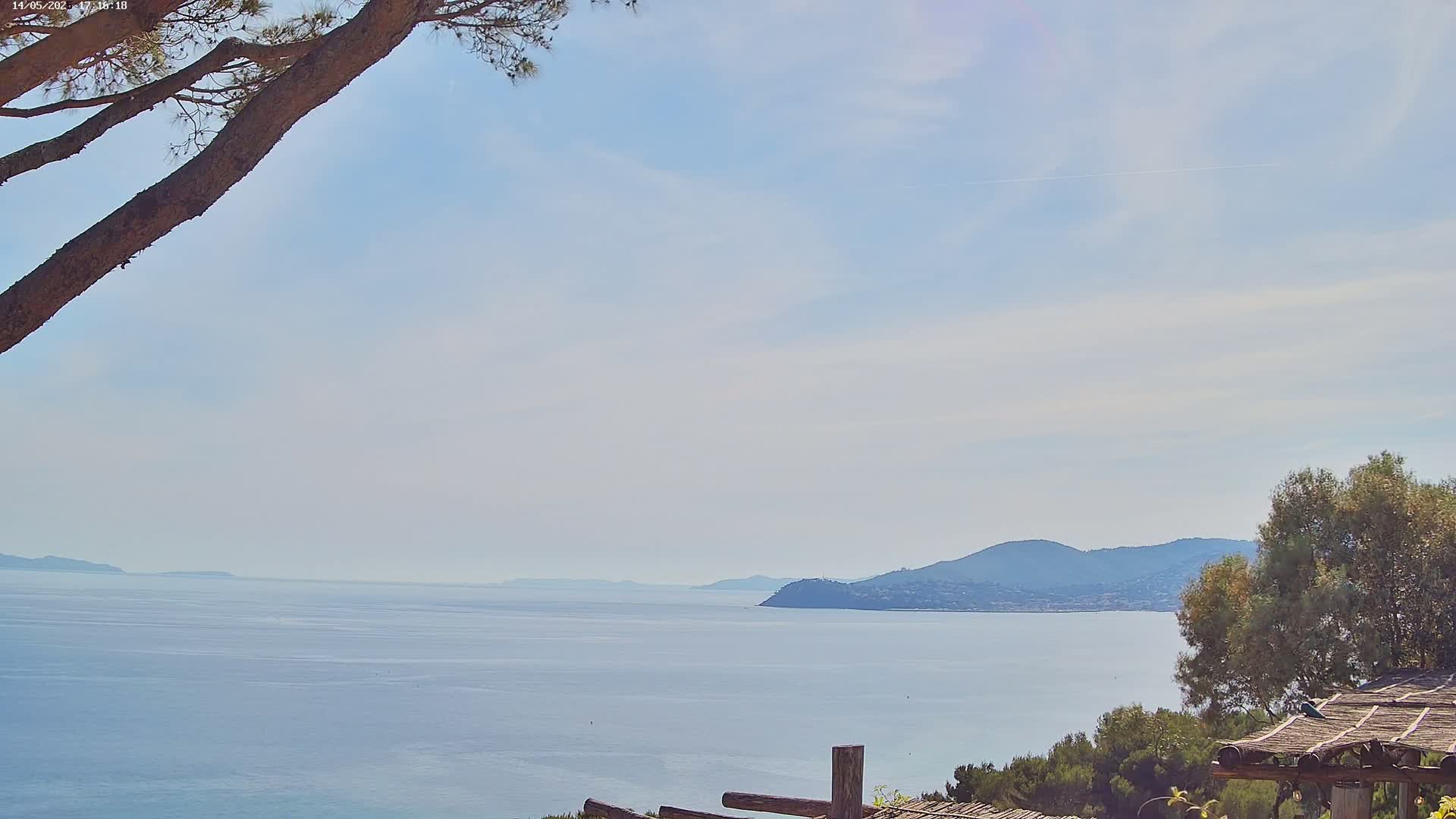 A tranquil ocean view on a partly cloudy day, seen from behind a tree branch and wooden structure, shows distant hills and vegetation.