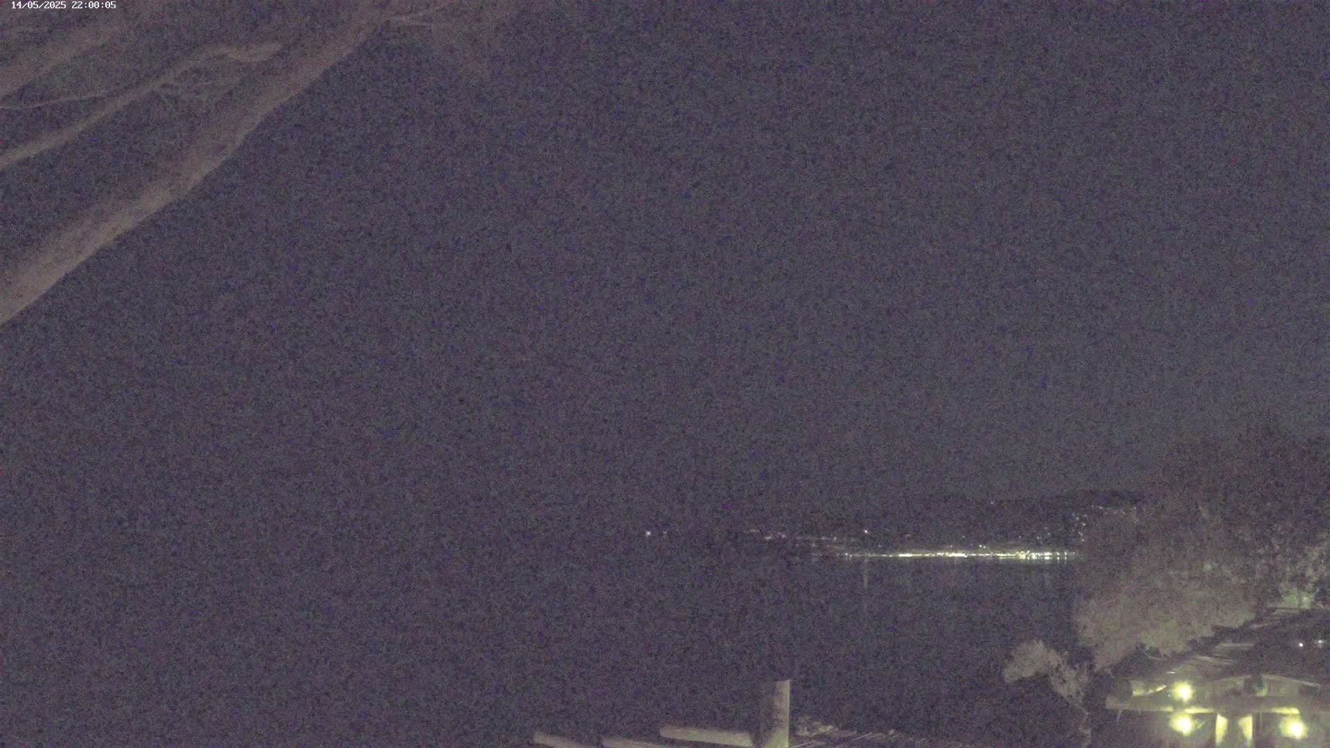 A nighttime, long-range view shows a dark body of water with faint lights on the far shore, and dimly lit buildings and trees in the foreground.