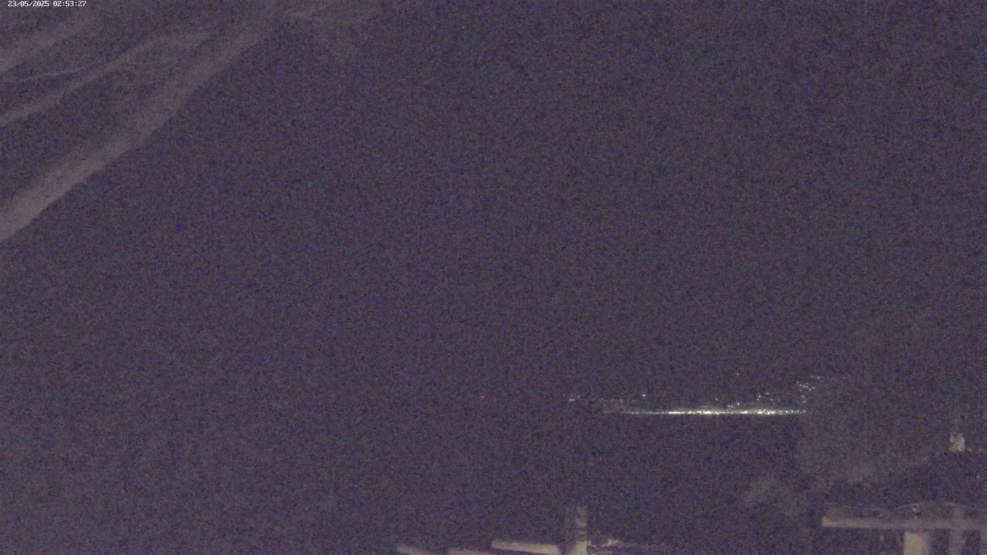 A nighttime long shot shows a line of distant lights across a dark body of water, partially obscured by shadows from overhanging tree branches.