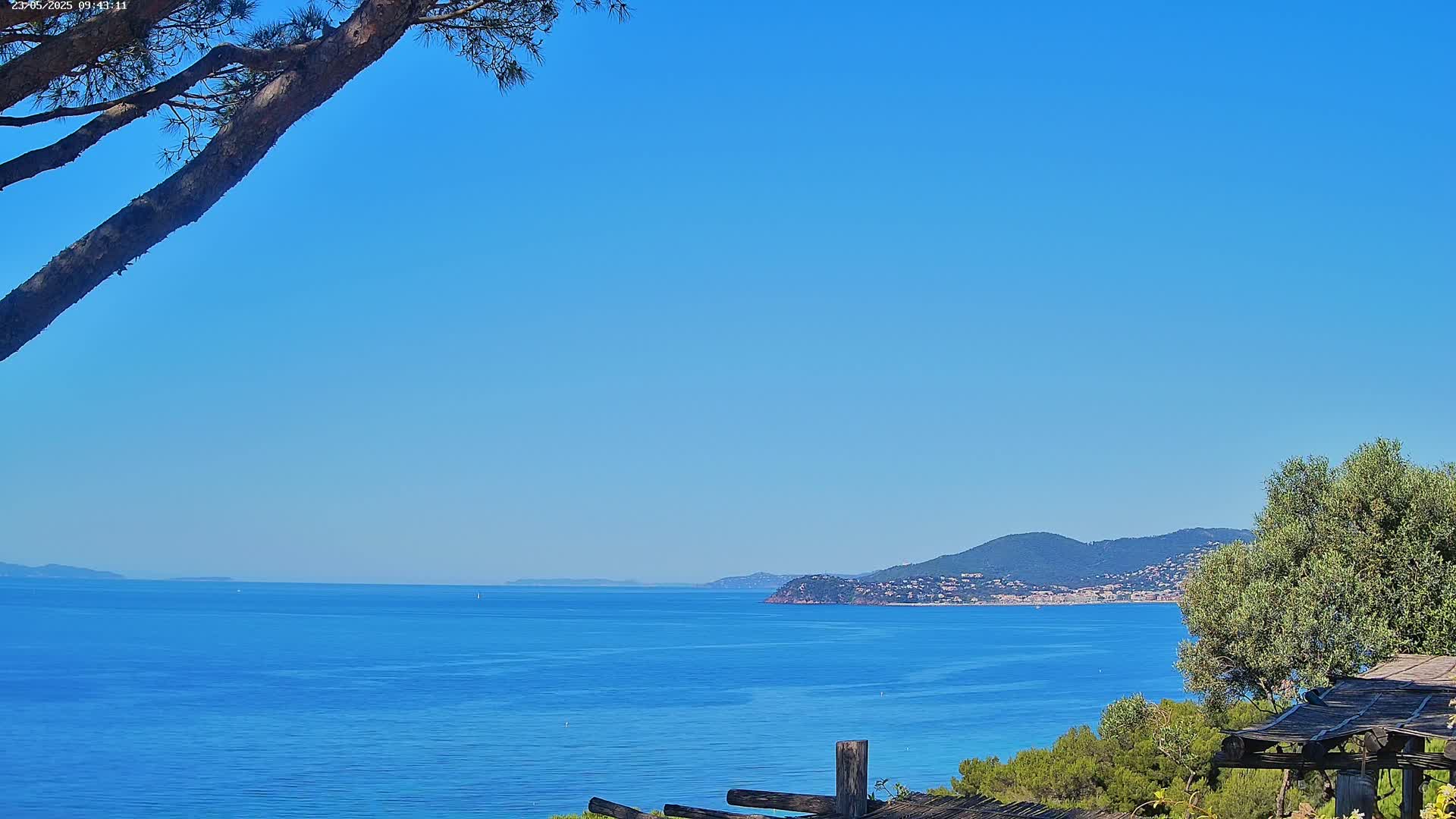 A tranquil ocean scene under a clear blue sky shows a calm sea, distant mountains, and a portion of a wooden structure in the foreground.