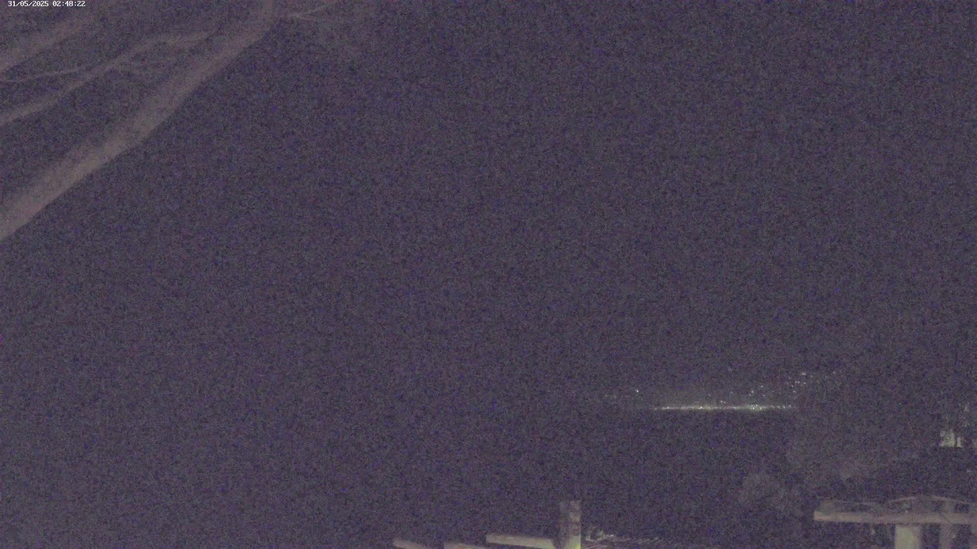A nighttime long shot shows a distant line of lights on the horizon under a dark, clear sky, with part of a tree branch in the upper left corner and what appears to be a wooden structure in the bottom right.