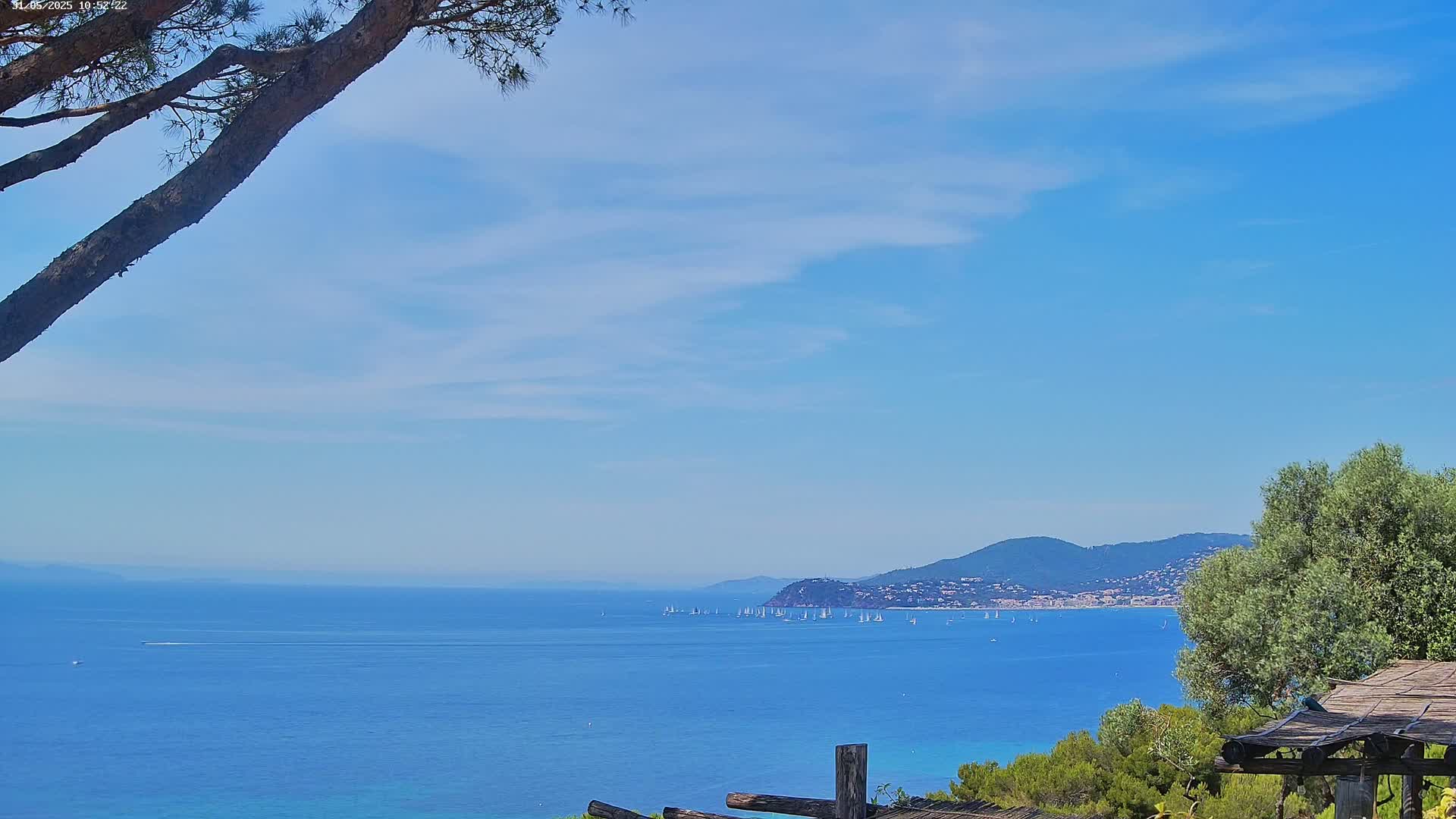 A partly cloudy blue sky overlooks a calm blue sea with numerous sailboats, a distant mountain range, and a portion of a wooden structure in the foreground.