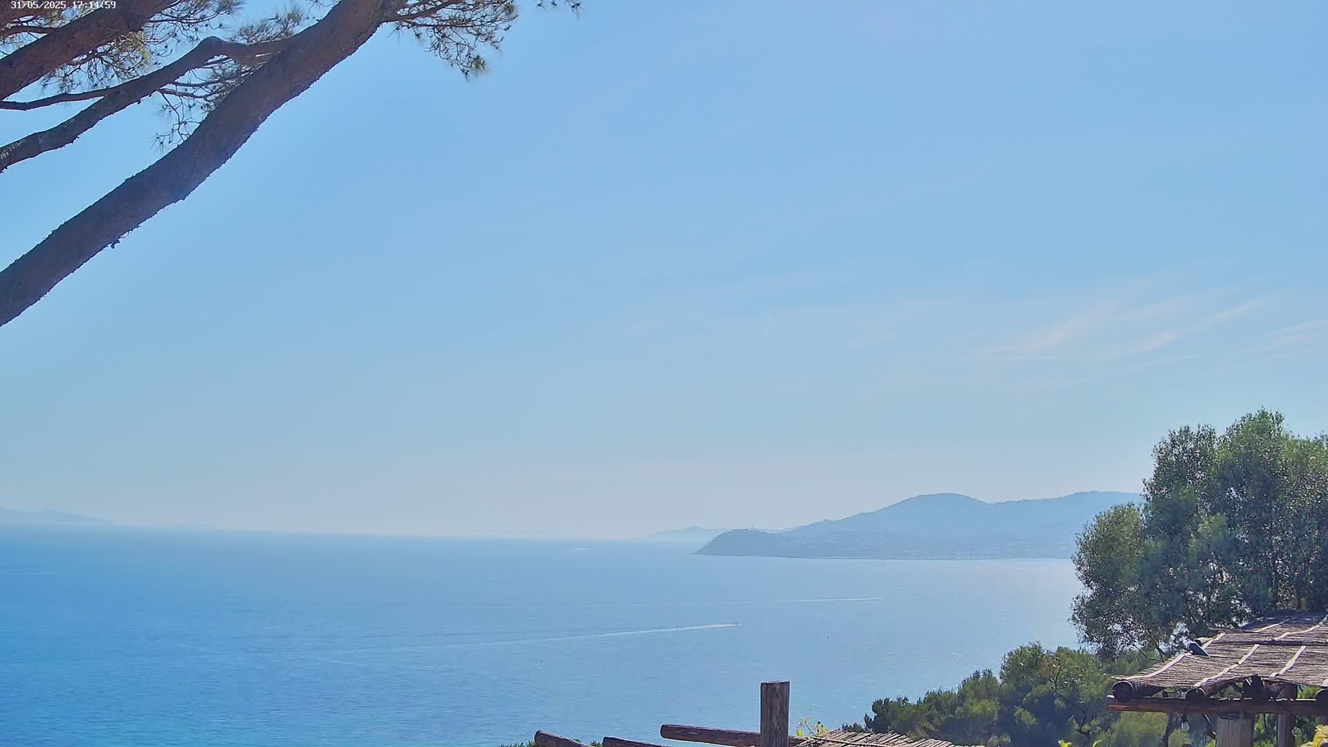 A calm, blue sea stretches to distant mountains under a clear, light-blue sky, viewed from a vantage point partially shaded by a tree and a wooden structure.