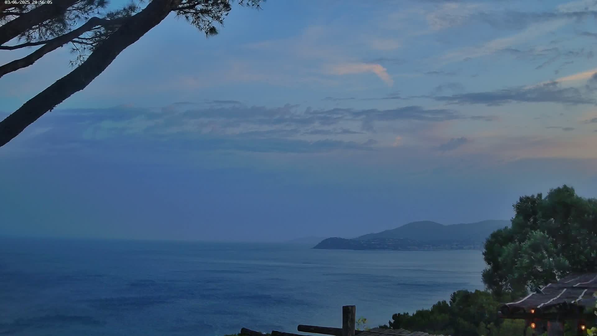 A calm, blue sea stretches to a distant, dark mountain under a partly cloudy twilight sky, viewed from a vantage point partially obscured by tree branches.